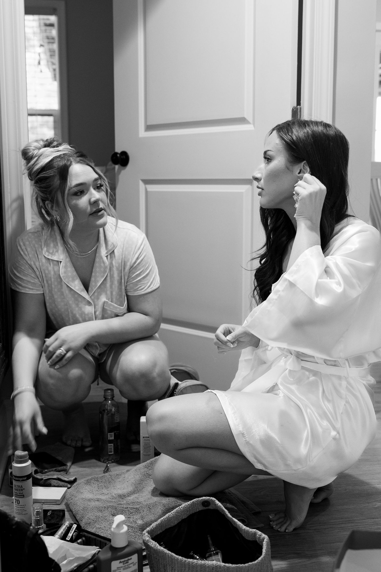 Two women getting ready, one in a white robe putting on earrings, the other in pajamas, sitting on the floor.