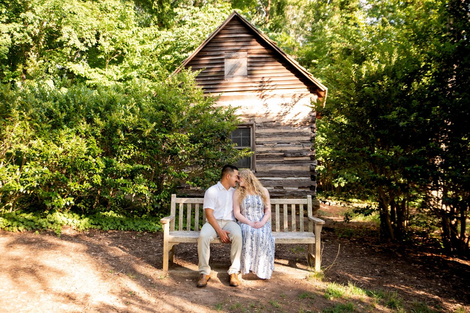 Couple sitting on a wooden bench in front of a rustic cabin surrounded by lush green trees.