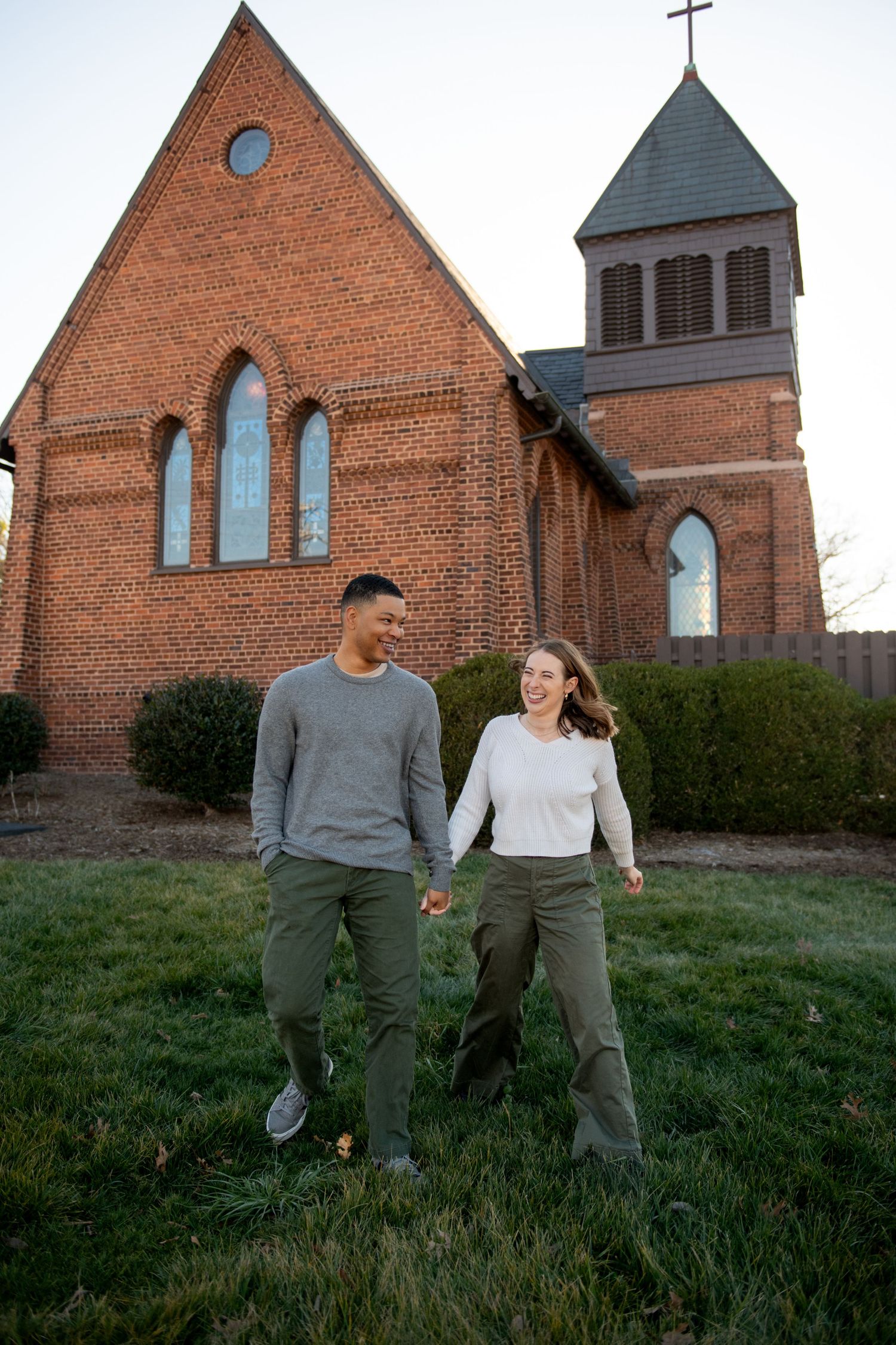 A couple in casual attire walks together on grass in front of a historic red brick church.