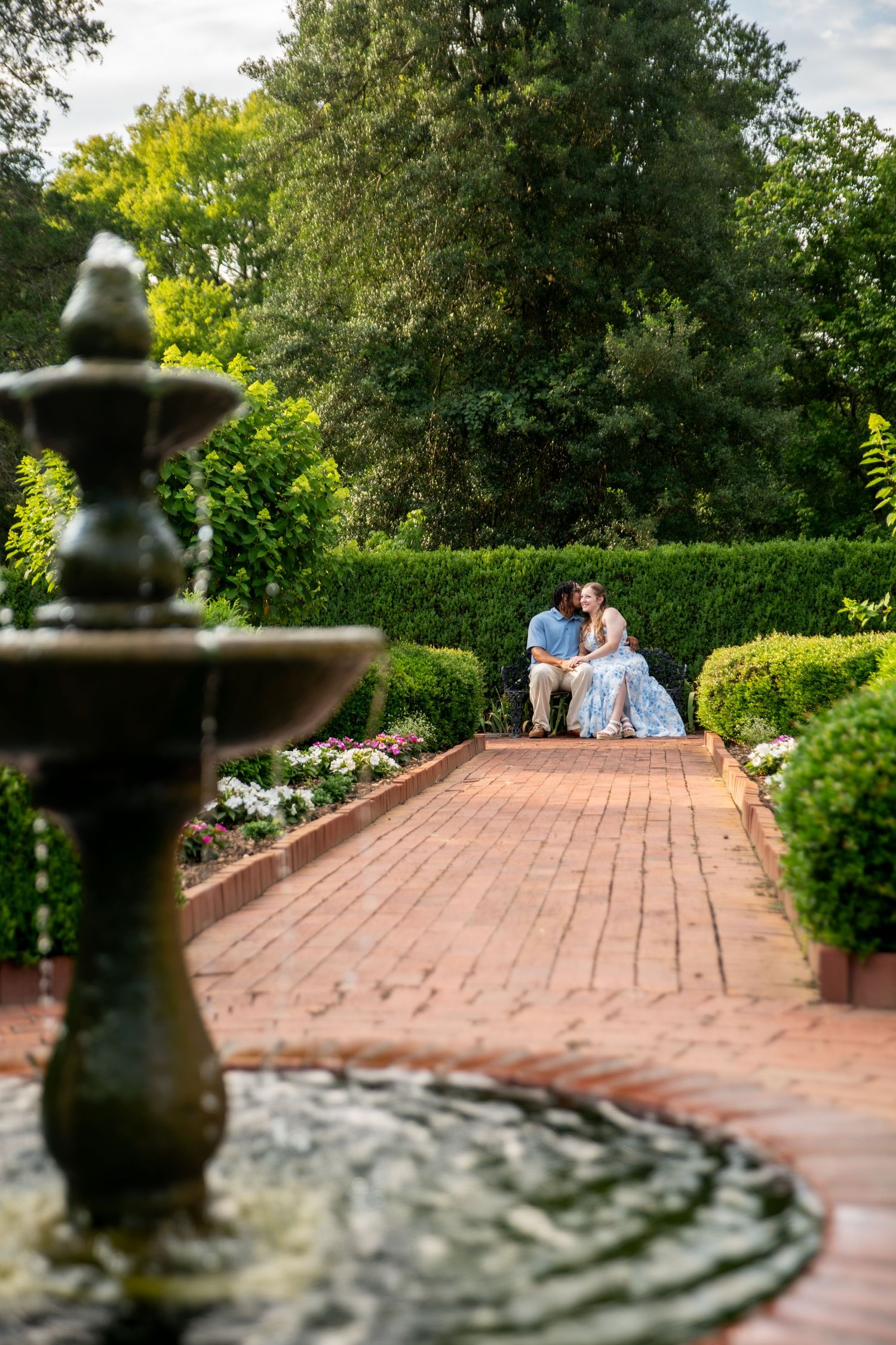 Couple sharing a romantic moment on a brick garden path with a fountain in the foreground and lush green hedges.