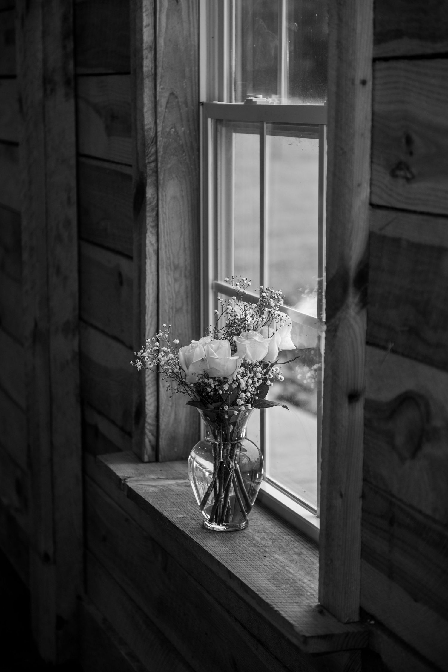 Black and white photo of a flower vase on a wooden windowsill in a rustic cabin interior.