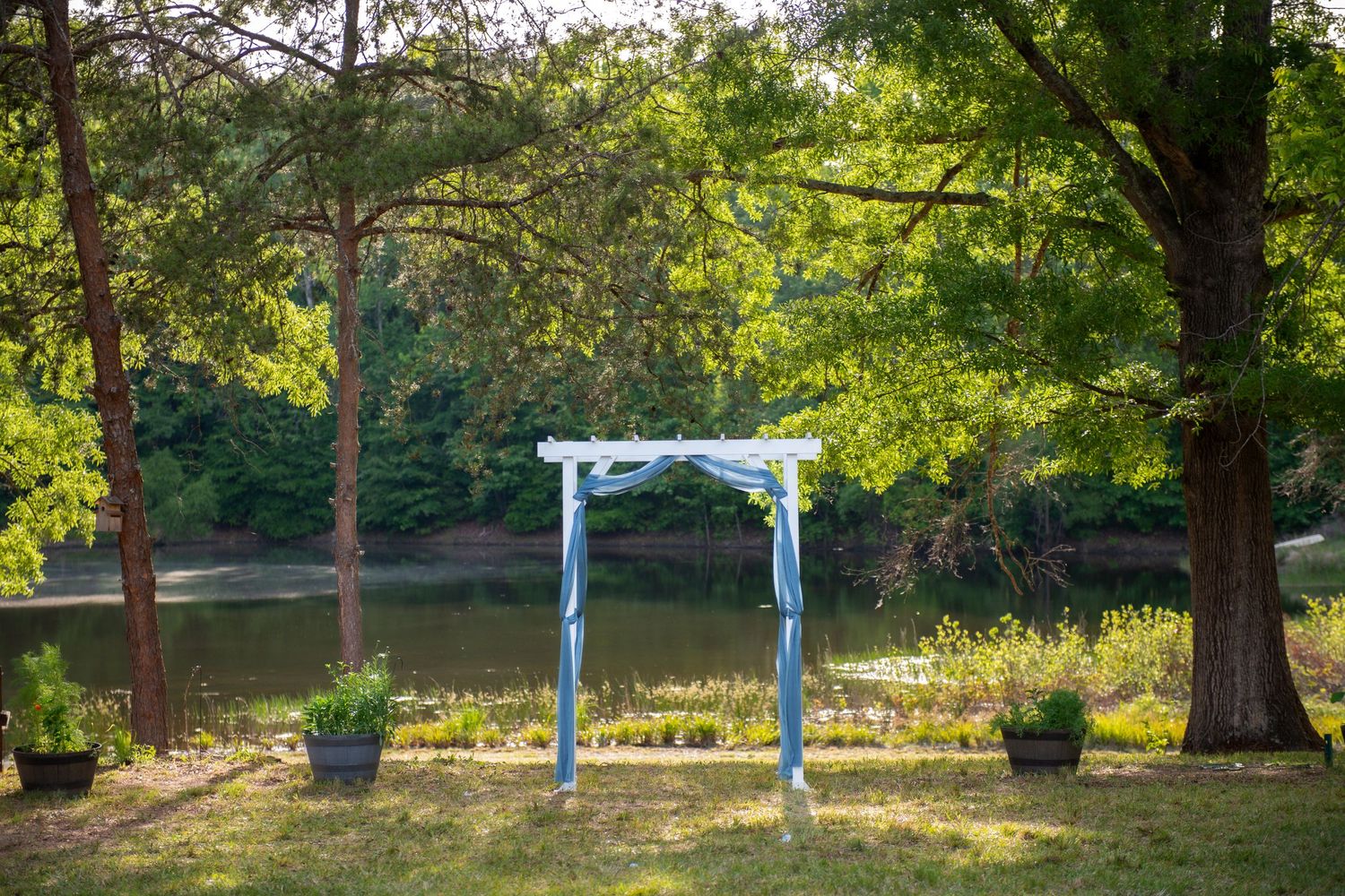 White wedding arch with blue ribbons set in a lush garden by a tranquil pond, surrounded by tall trees.