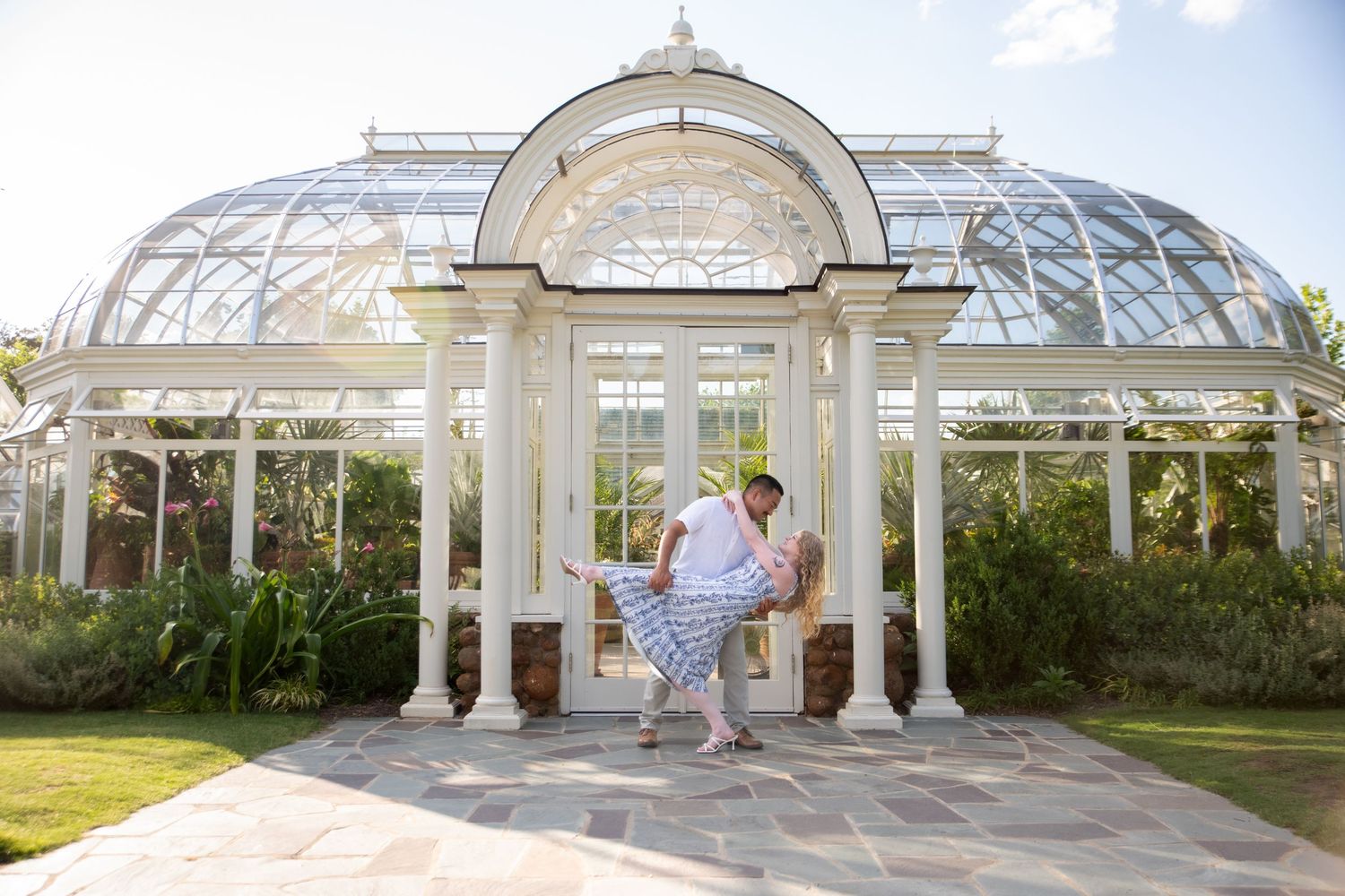 Groom dips bride in blue dress in romantic pose before elegant white glass greenhouse conservatory.