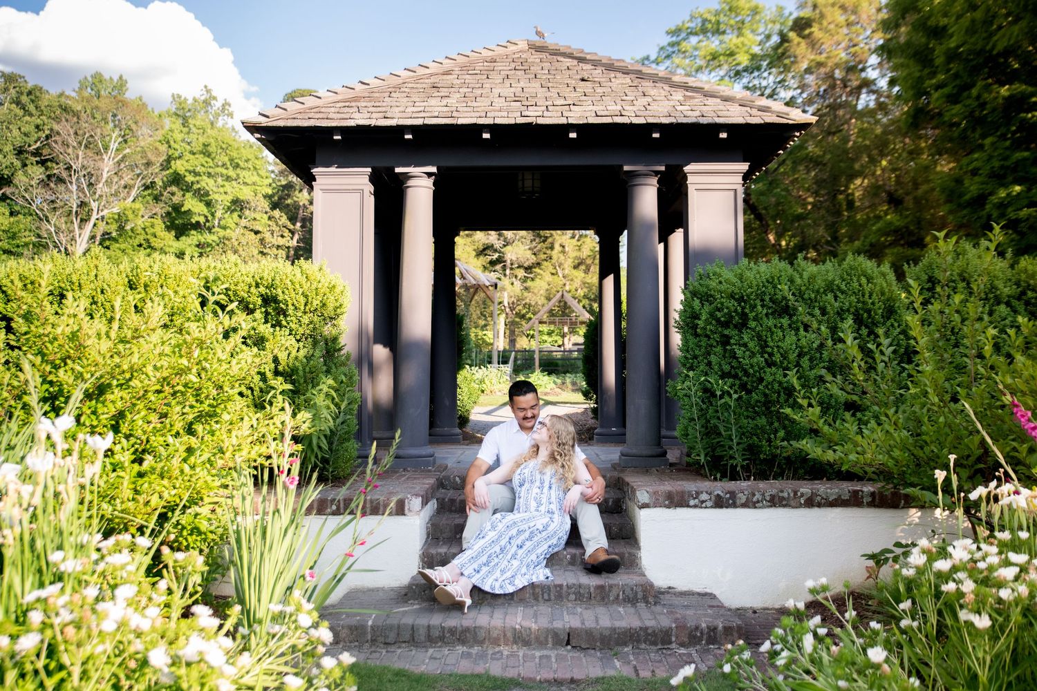Couple sitting on steps of elegant garden gazebo with dark columns, surrounded by lush greenery and flowers on a sunny day.