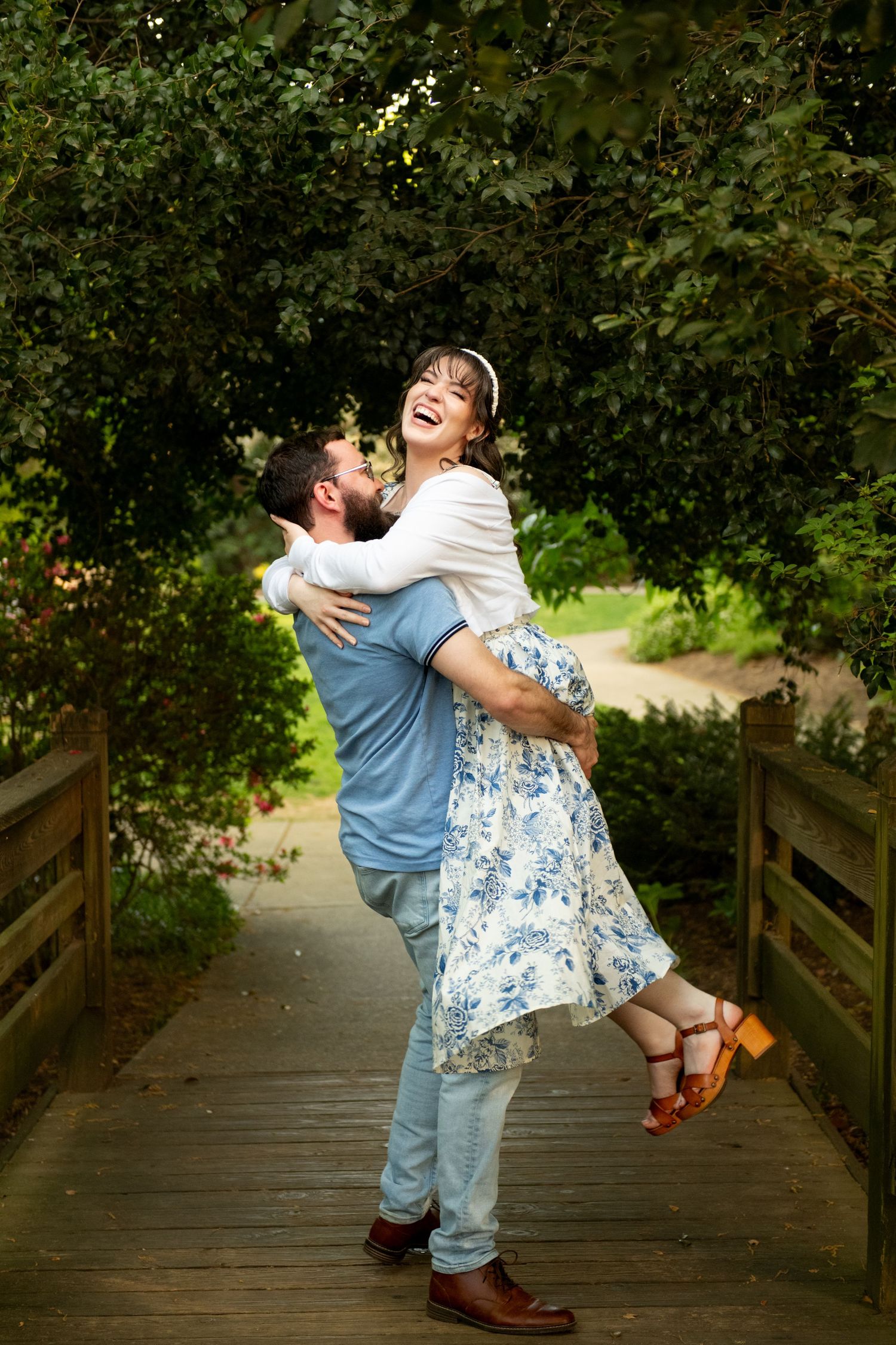 A joyful couple embraces on a wooden bridge surrounded by lush green trees, as the man lifts the laughing woman.