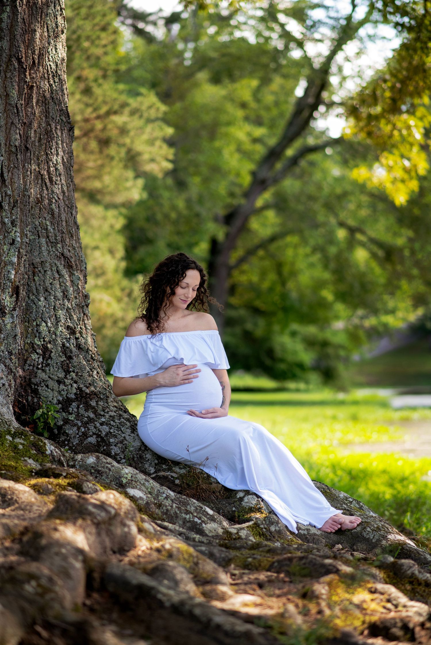 A person in a white dress rests peacefully against a tree trunk in a sunlit park setting.