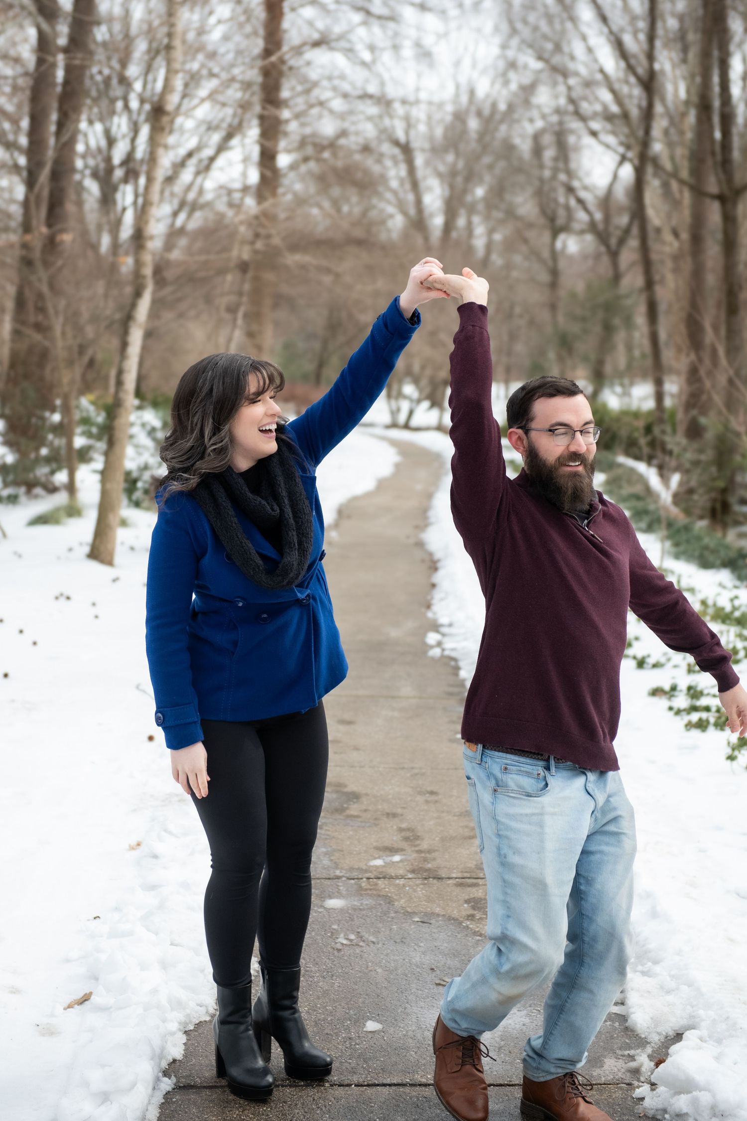 A playful couple dances and twirls together on a snow-covered path through winter trees.