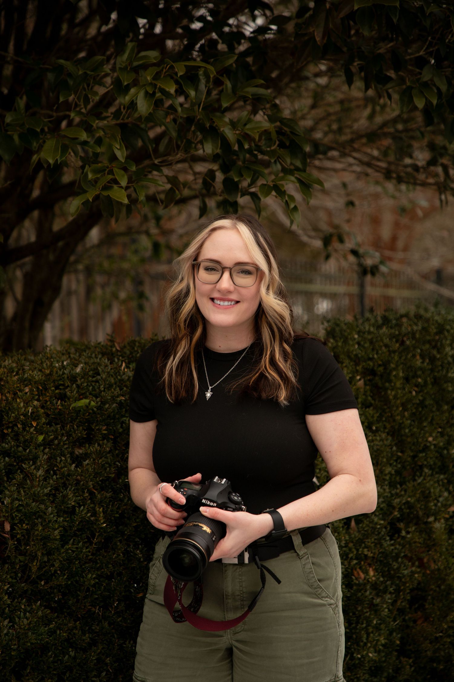 A photographer in glasses and casual attire holds a professional camera while posing outdoors near green hedges.