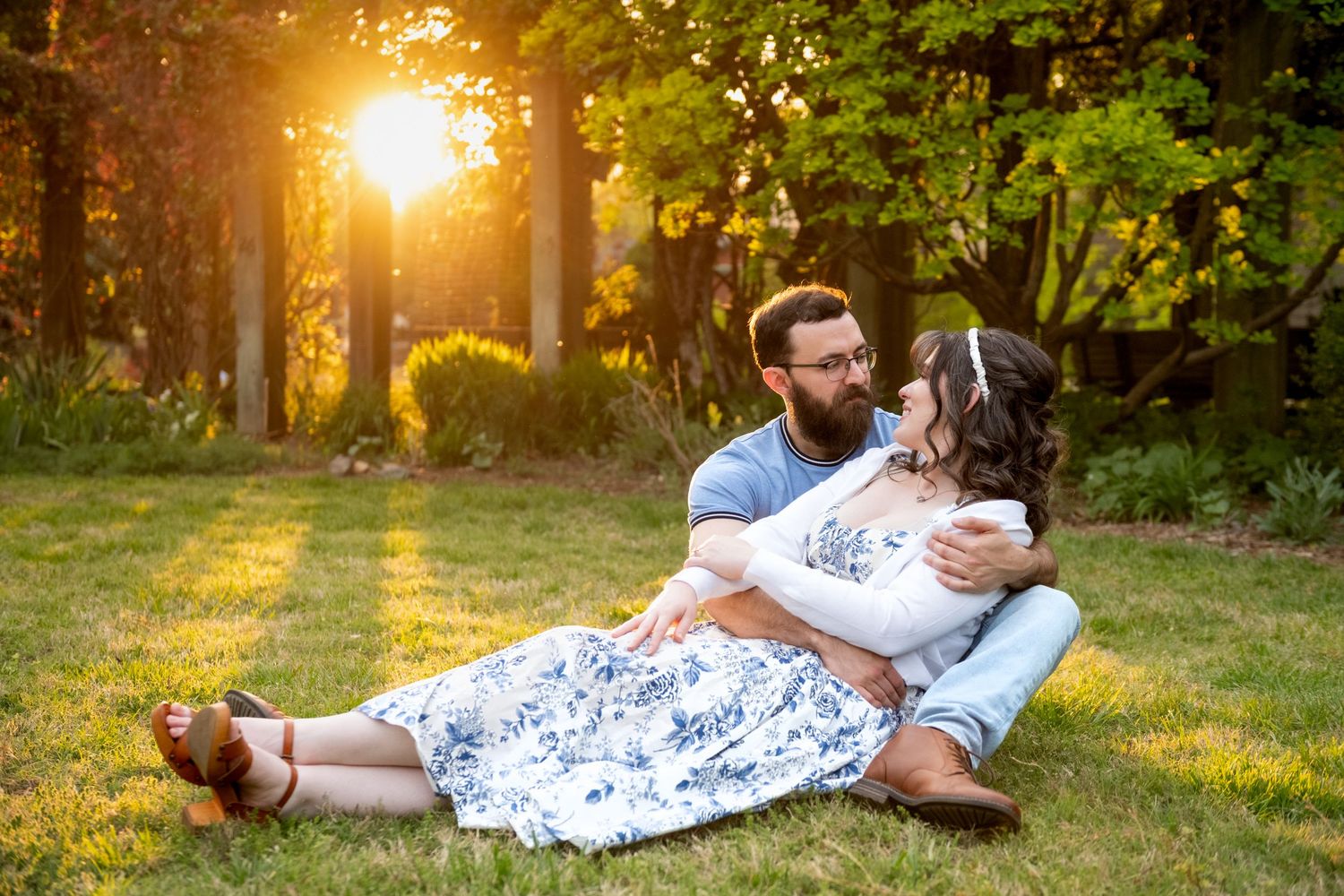 Couple sitting on green grass at golden hour sunset, man with beard embracing woman in floral skirt in a lush garden.