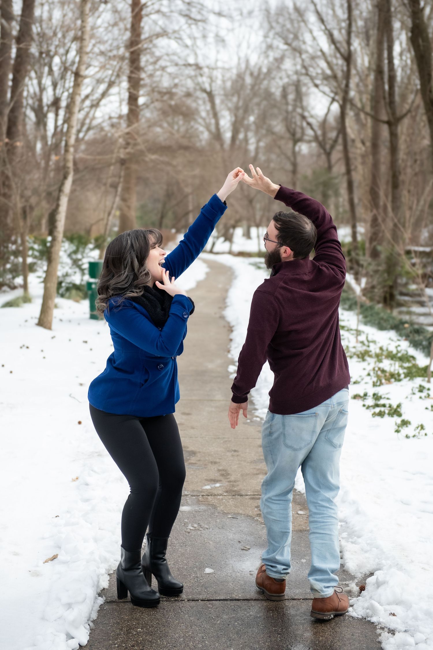 A couple performs joyful dance moves together on a snow-lined walkway in winter.