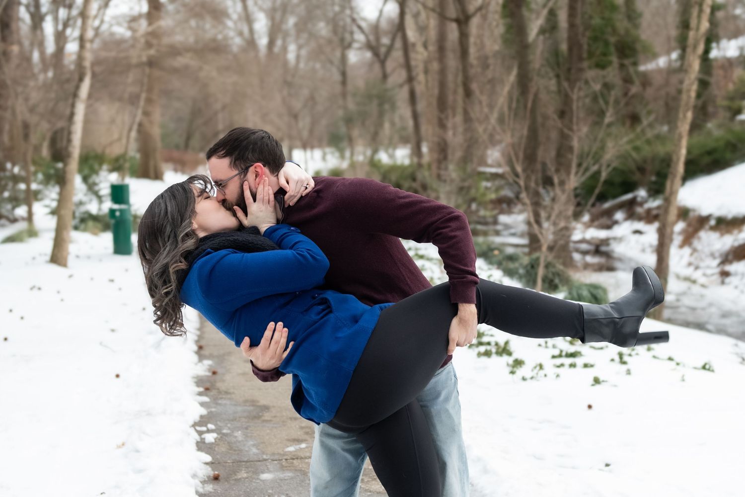 Two people embrace in a romantic dance pose against a snowy winter landscape with bare trees in the background.