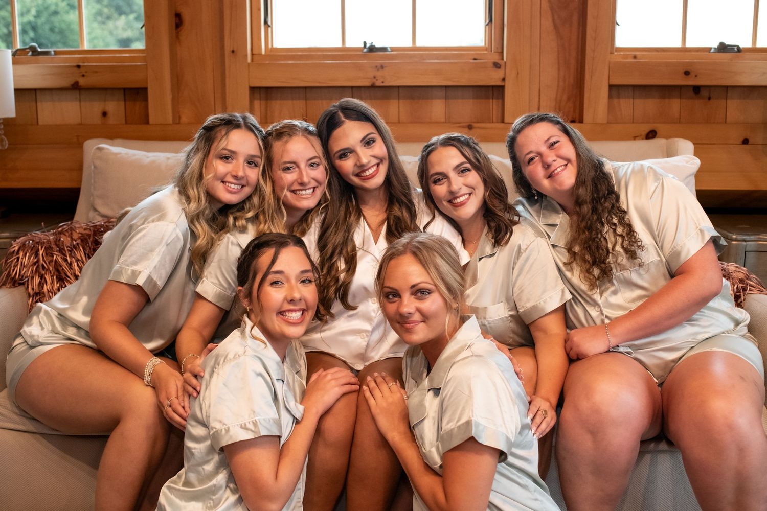 Seven young women in matching white button-up shirts smiling together on a couch in a cozy wood-paneled room.