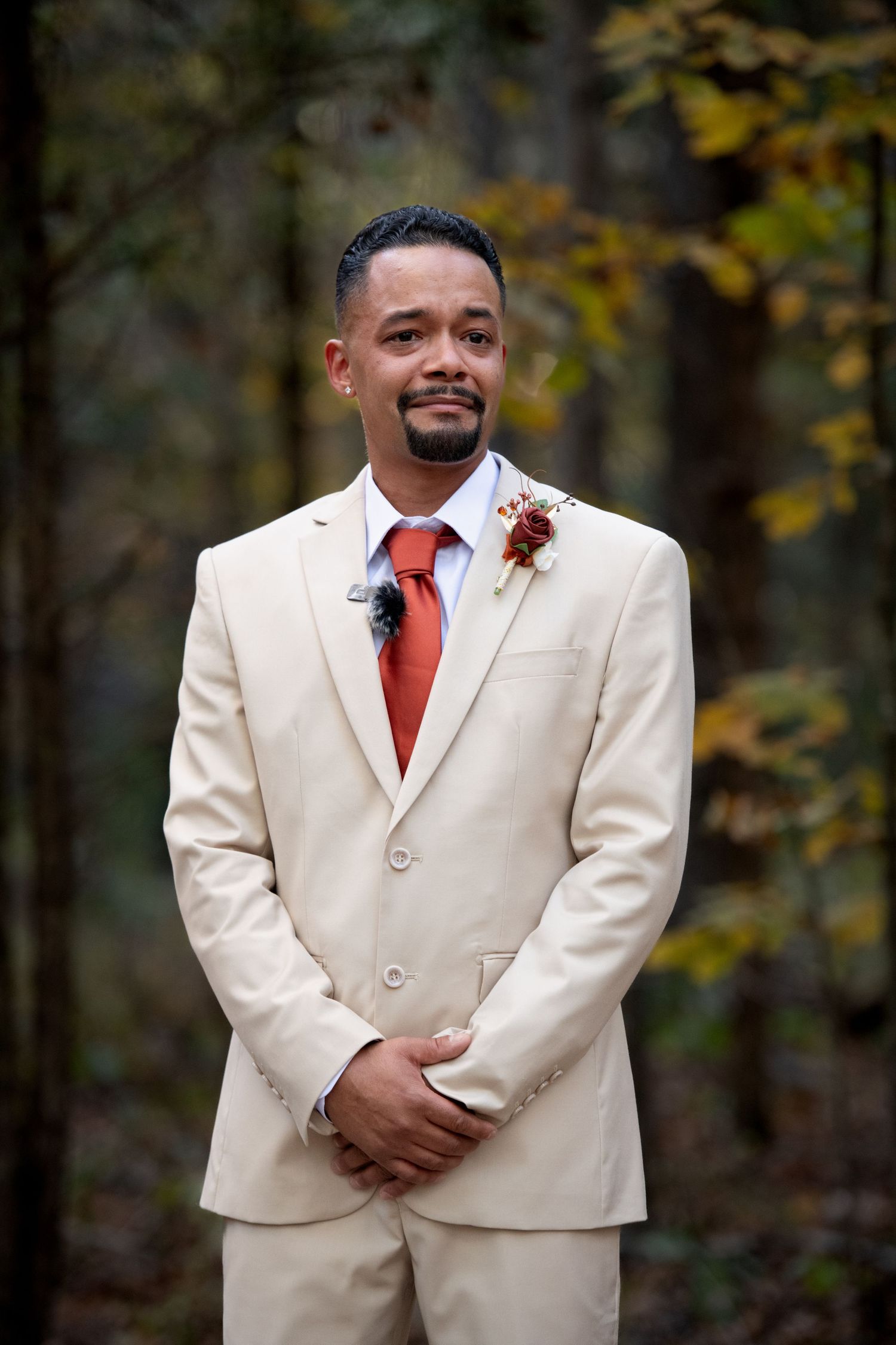 Groom in cream suit with red tie and boutonniere smiling outdoors in autumn forest setting.