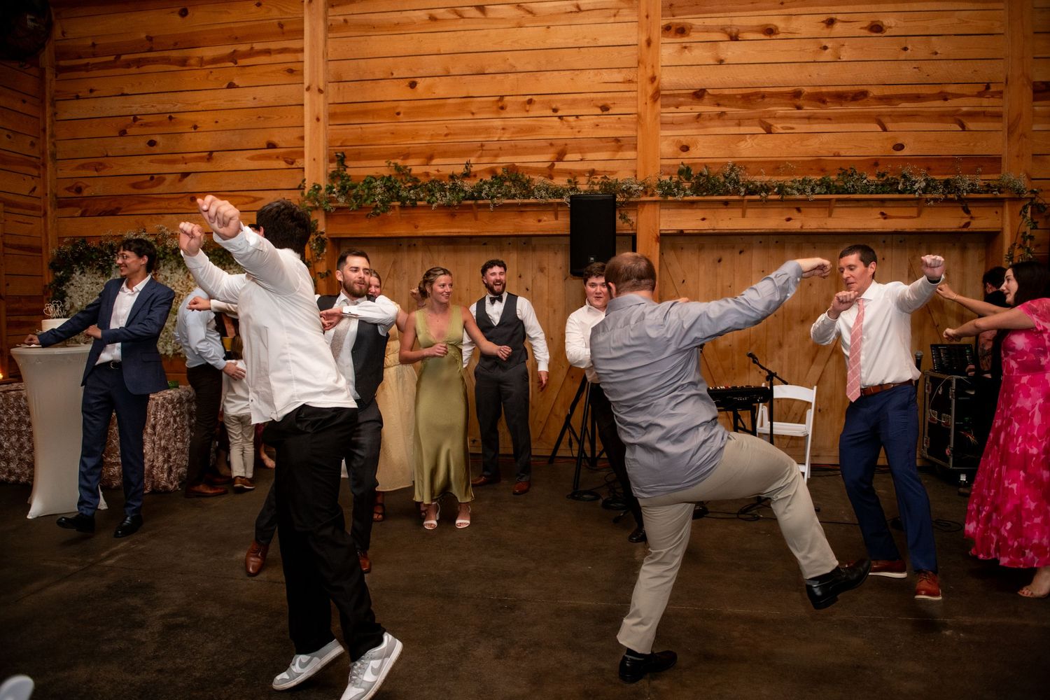 Guests dancing energetically at a rustic barn wedding reception with wood-paneled walls and greenery decorations.