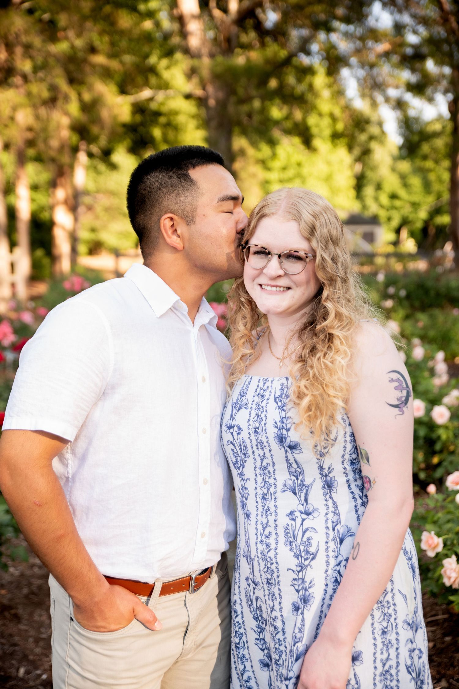 A man in a white shirt kisses a smiling blonde woman in a blue floral dress outdoors in a garden.