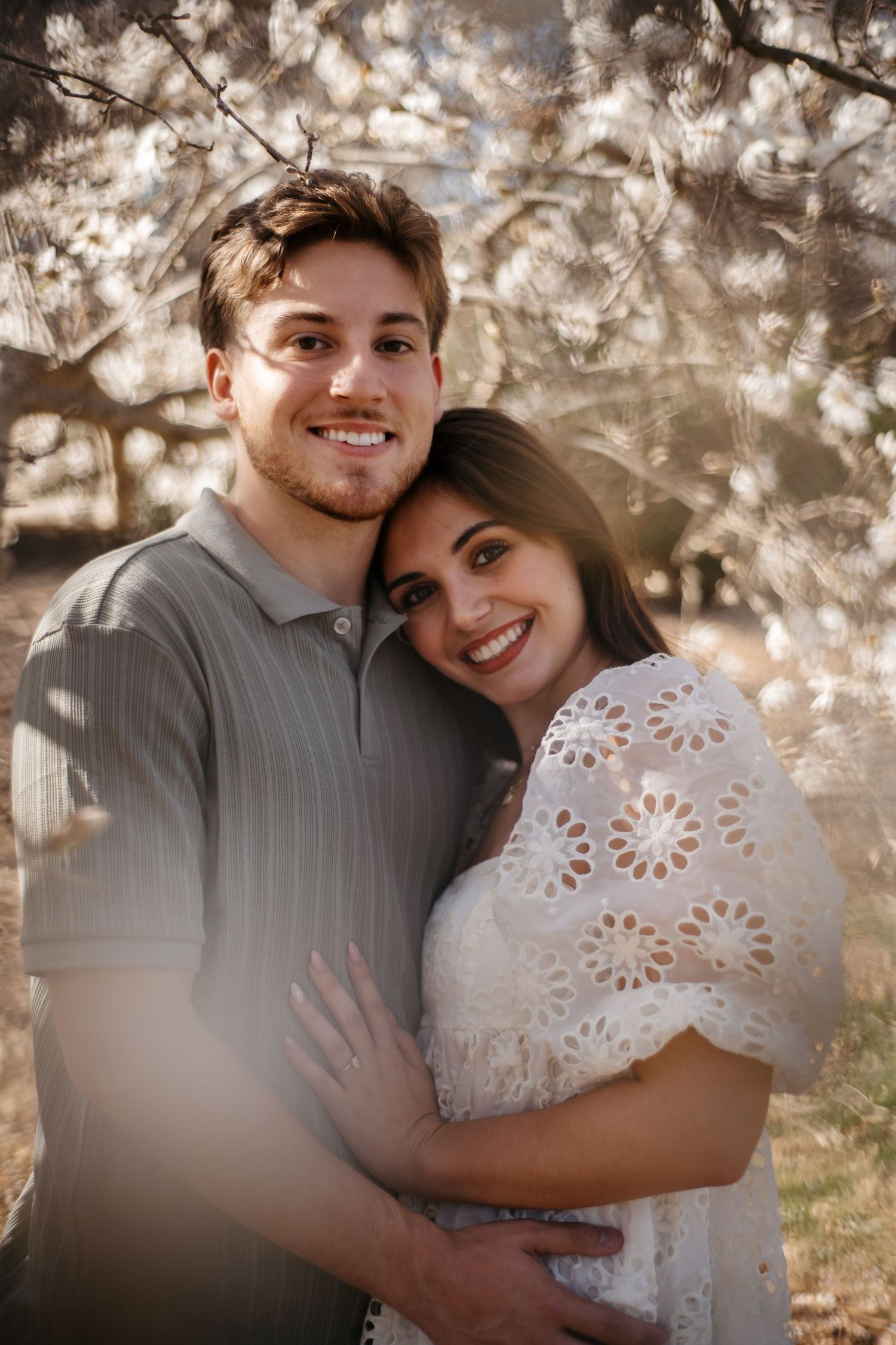 A romantic couple embraces under blooming spring trees with soft bokeh lighting in the background.