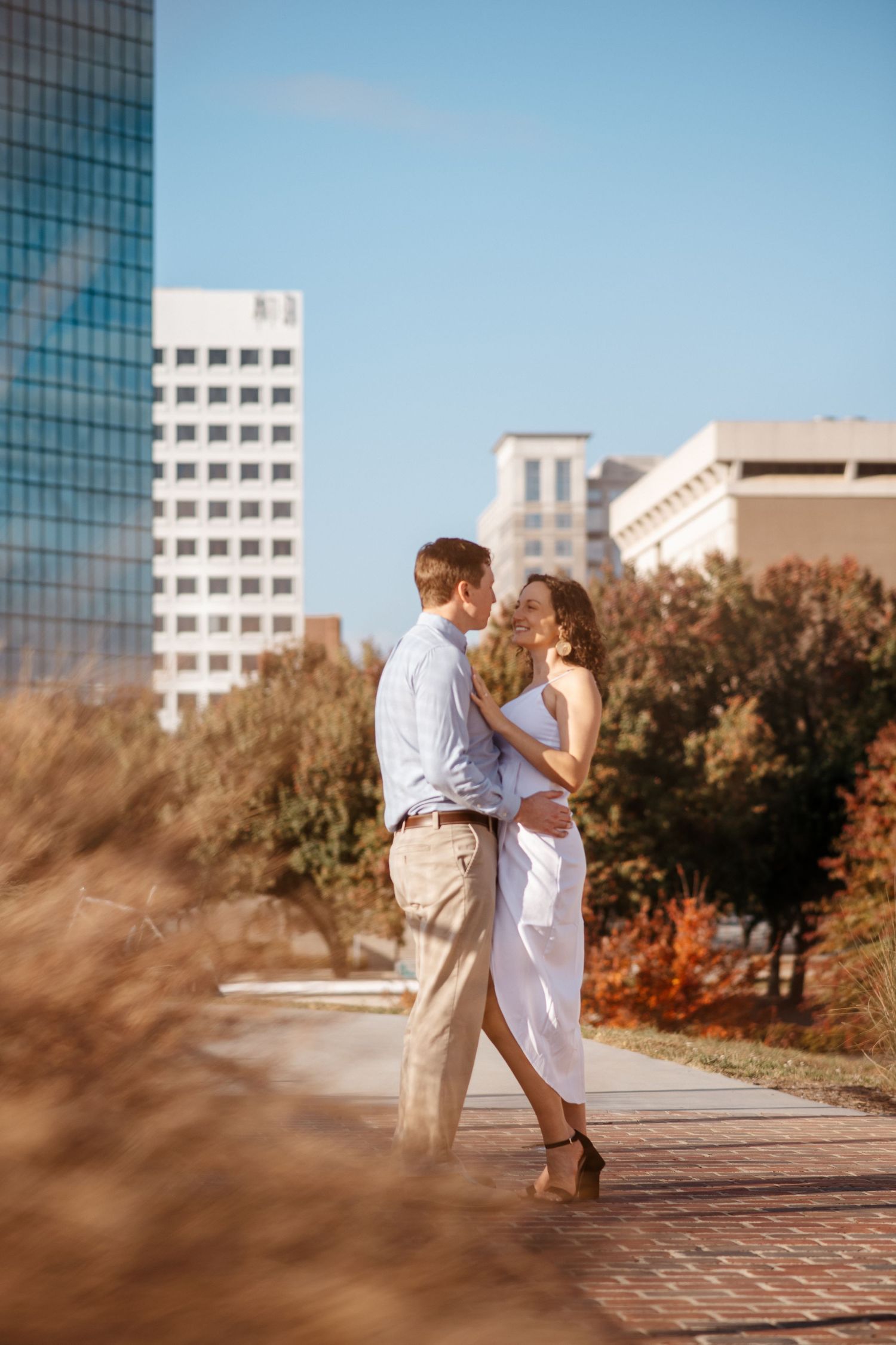 A romantic couple embracing on an urban pathway with fall foliage and buildings in background.