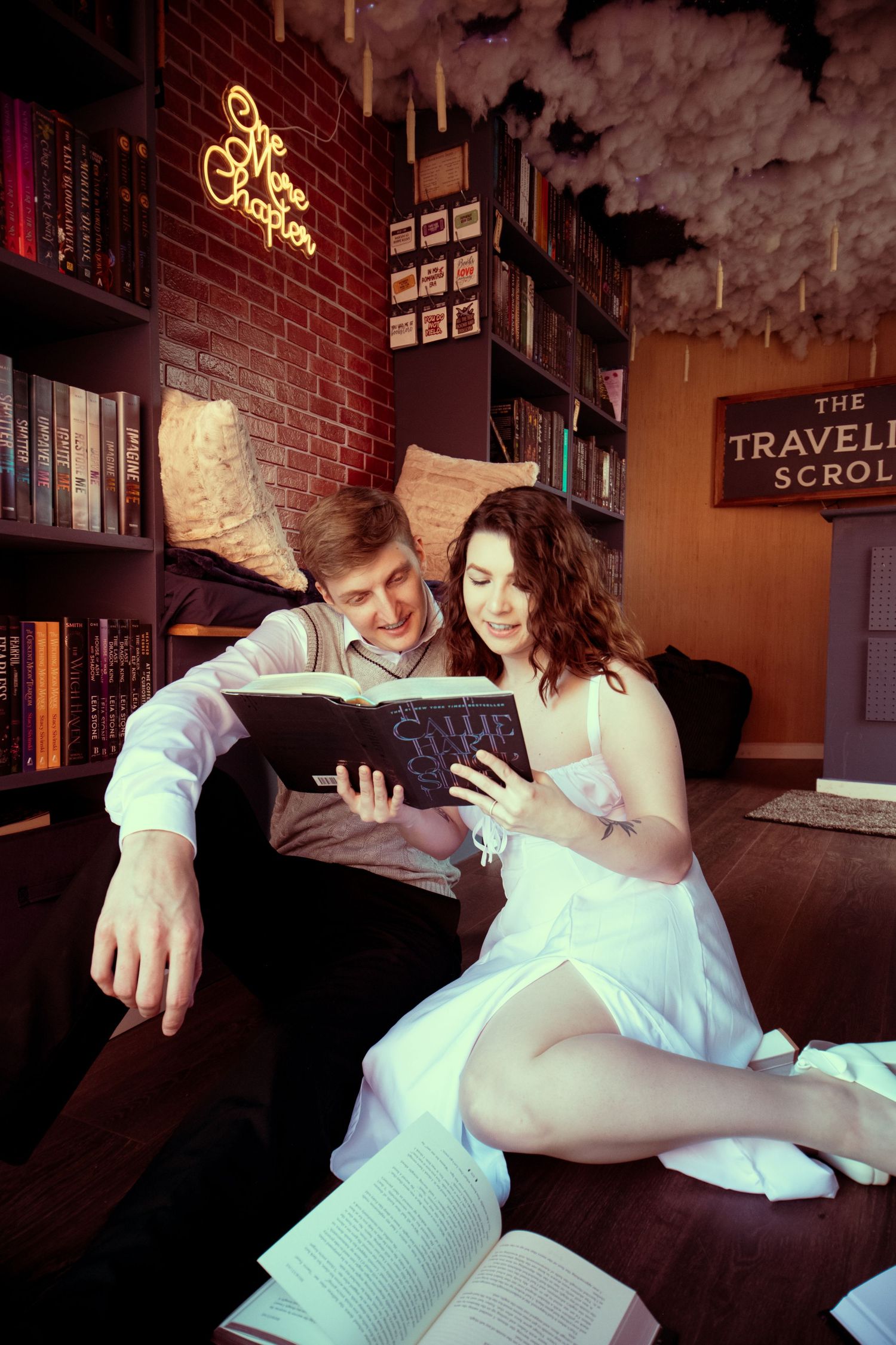A couple sits on the floor reading a book together in a cozy library with bookshelves and neon sign decor.