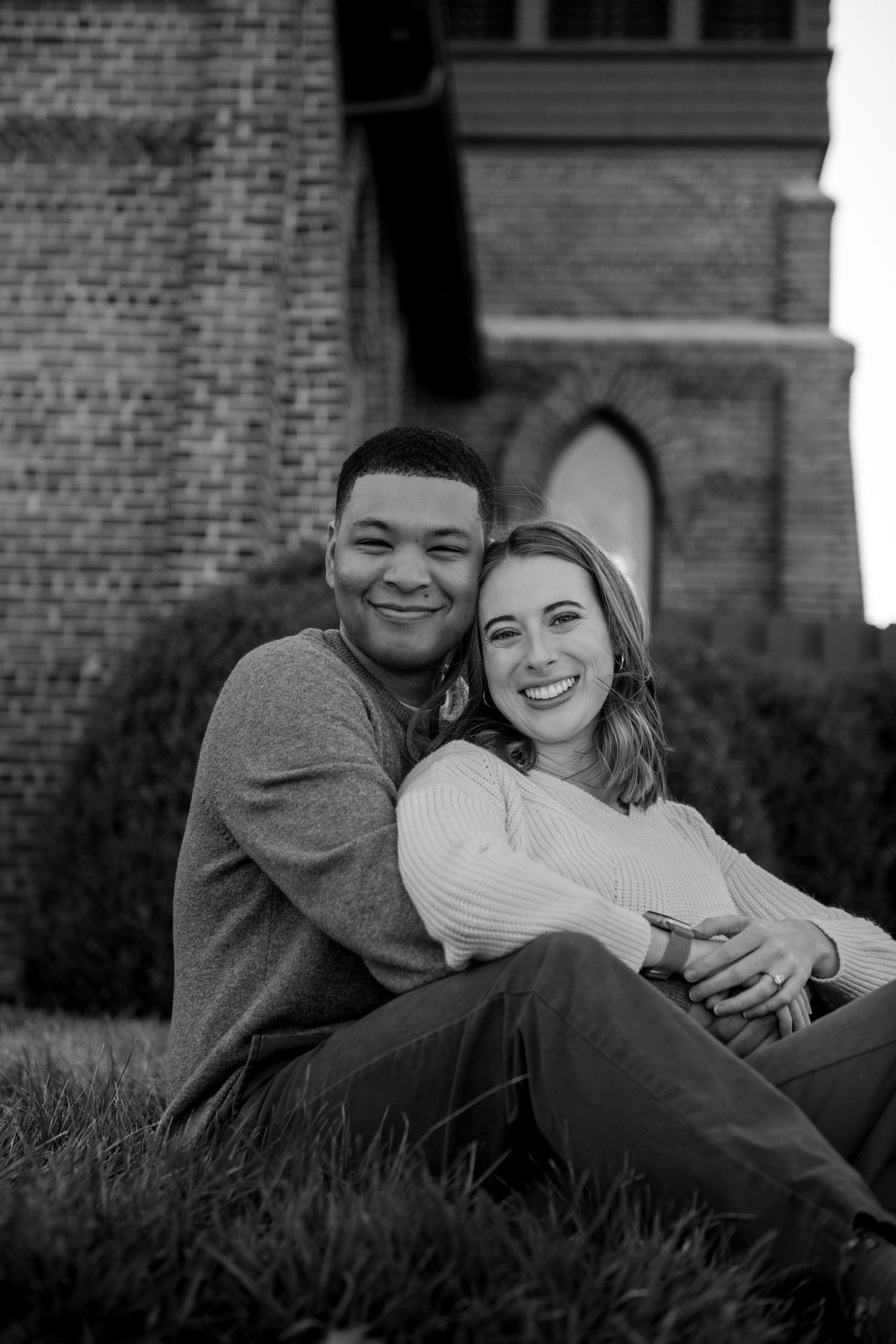 A couple sits together on grass near a brick building in a black and white portrait.