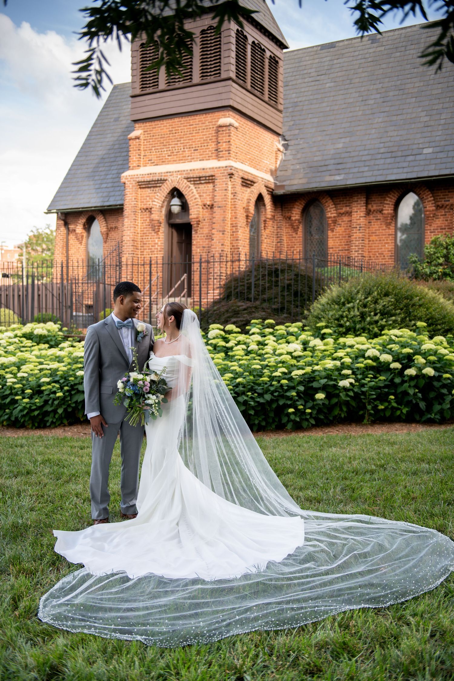 Bride and groom stand together on grass before a red brick church, her long cathedral veil flowing behind them.