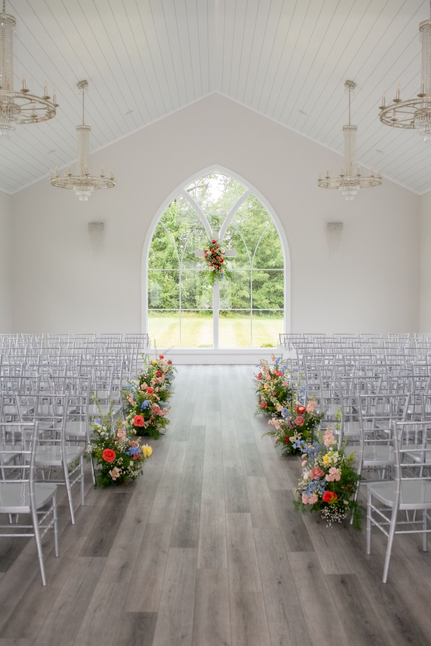 Elegant white wedding chapel with floral aisle arrangements, ghost chairs, arched window, and crystal chandeliers.