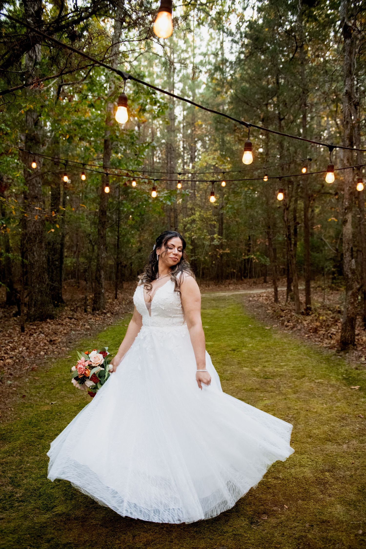 String lights illuminate a wooded outdoor setting with a white lace wedding dress twirling in motion.