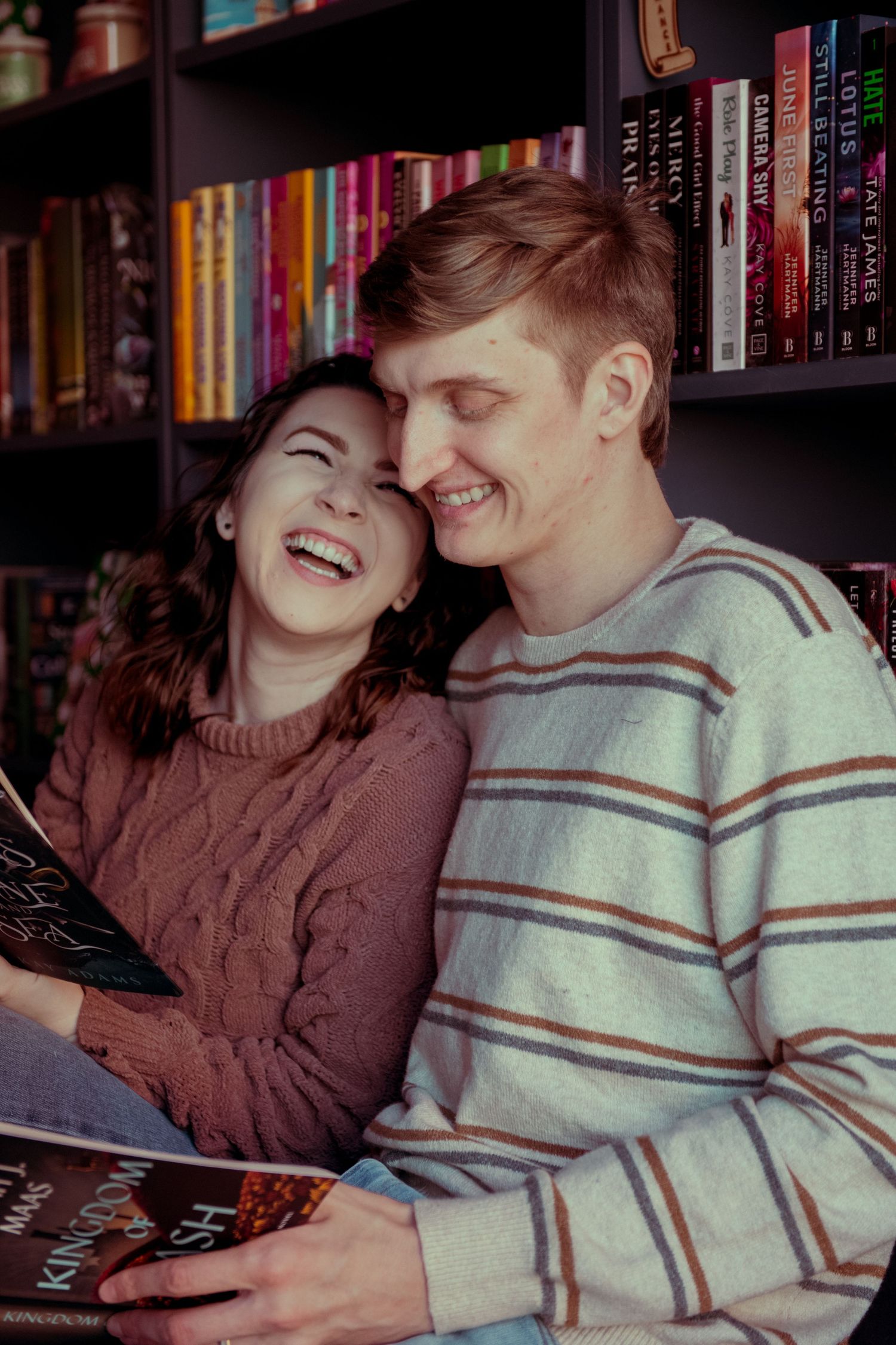 A happy couple laughs together while reading a book in front of a colorful bookshelf.