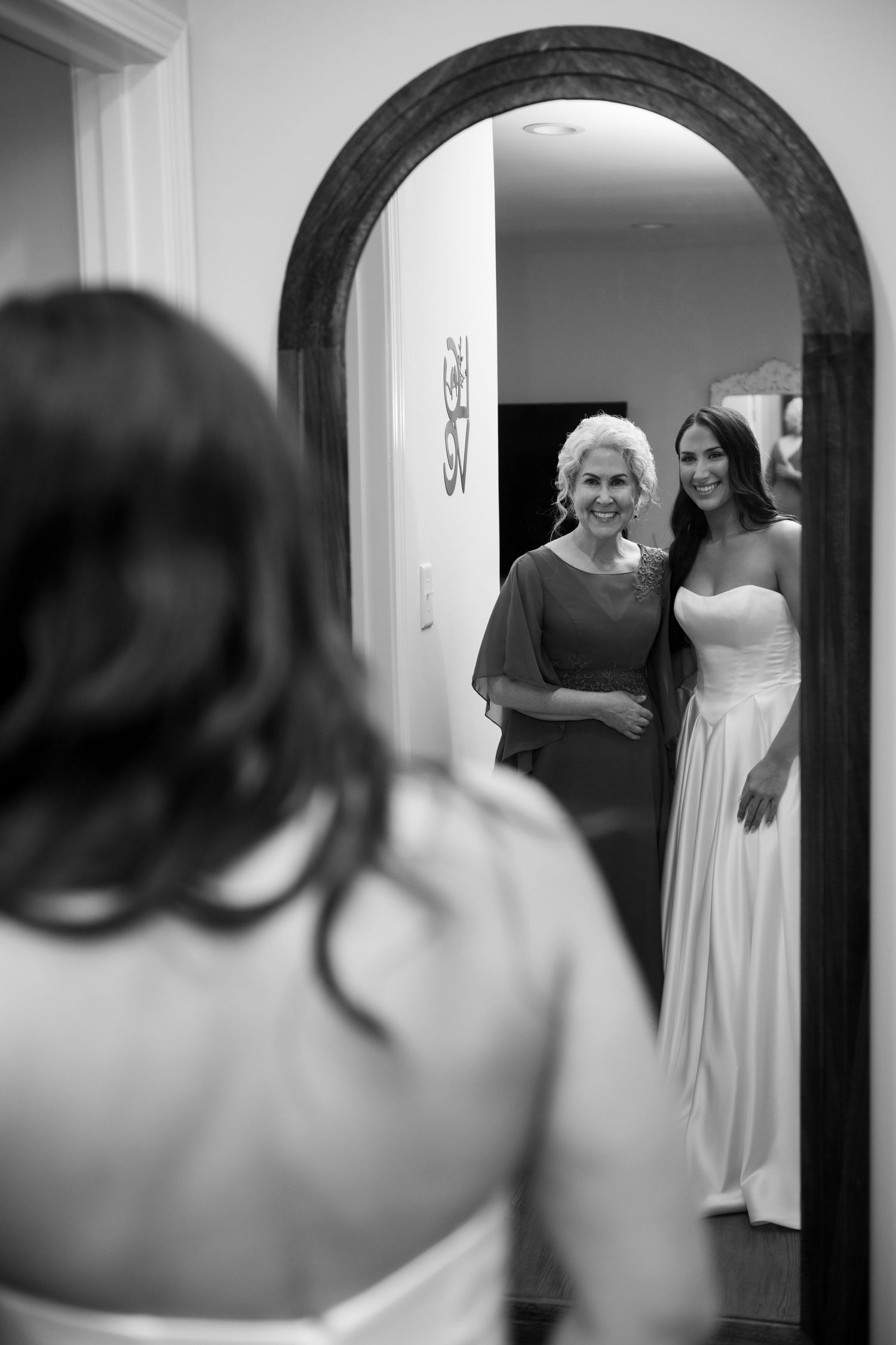 Black and white photo of a bride in a white gown posing with an older woman in a mirror reflection.
