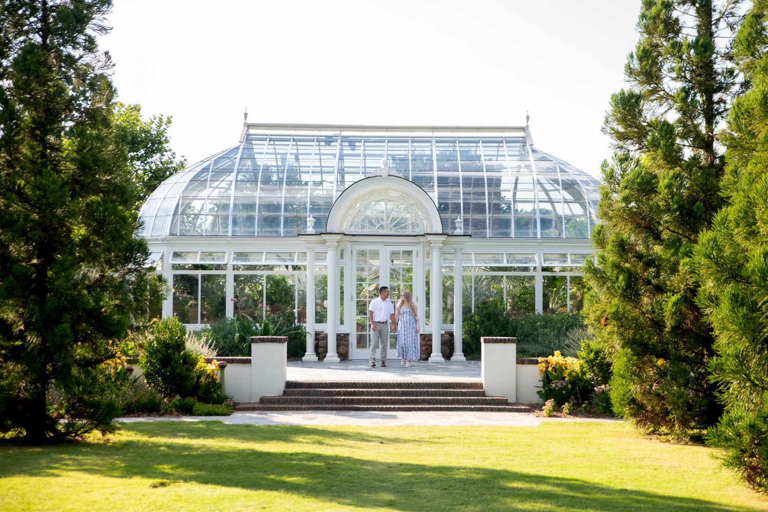 Couple standing in front of an elegant white glass greenhouse conservatory surrounded by lush green trees and lawn.