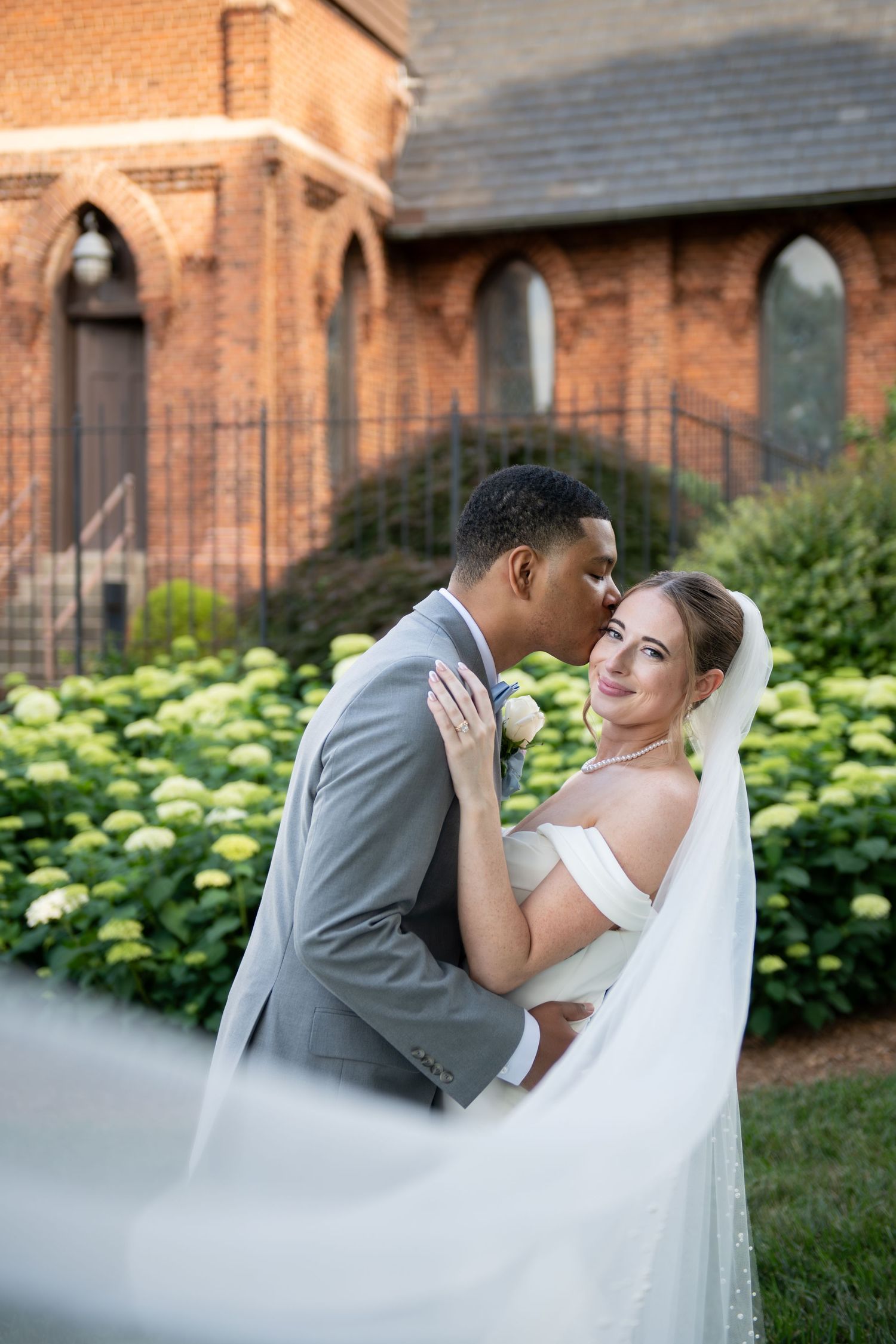 Groom in gray suit kisses bride in white off-shoulder gown near brick church with hydrangeas.