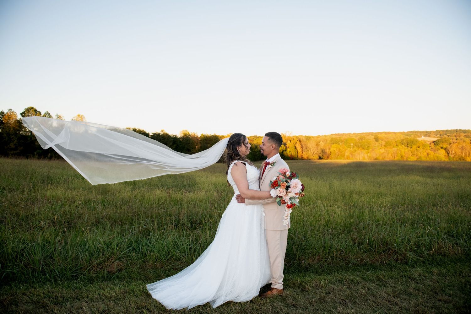 A flowing white veil billows in the breeze as a couple embraces in a sunlit meadow during their wedding photoshoot.