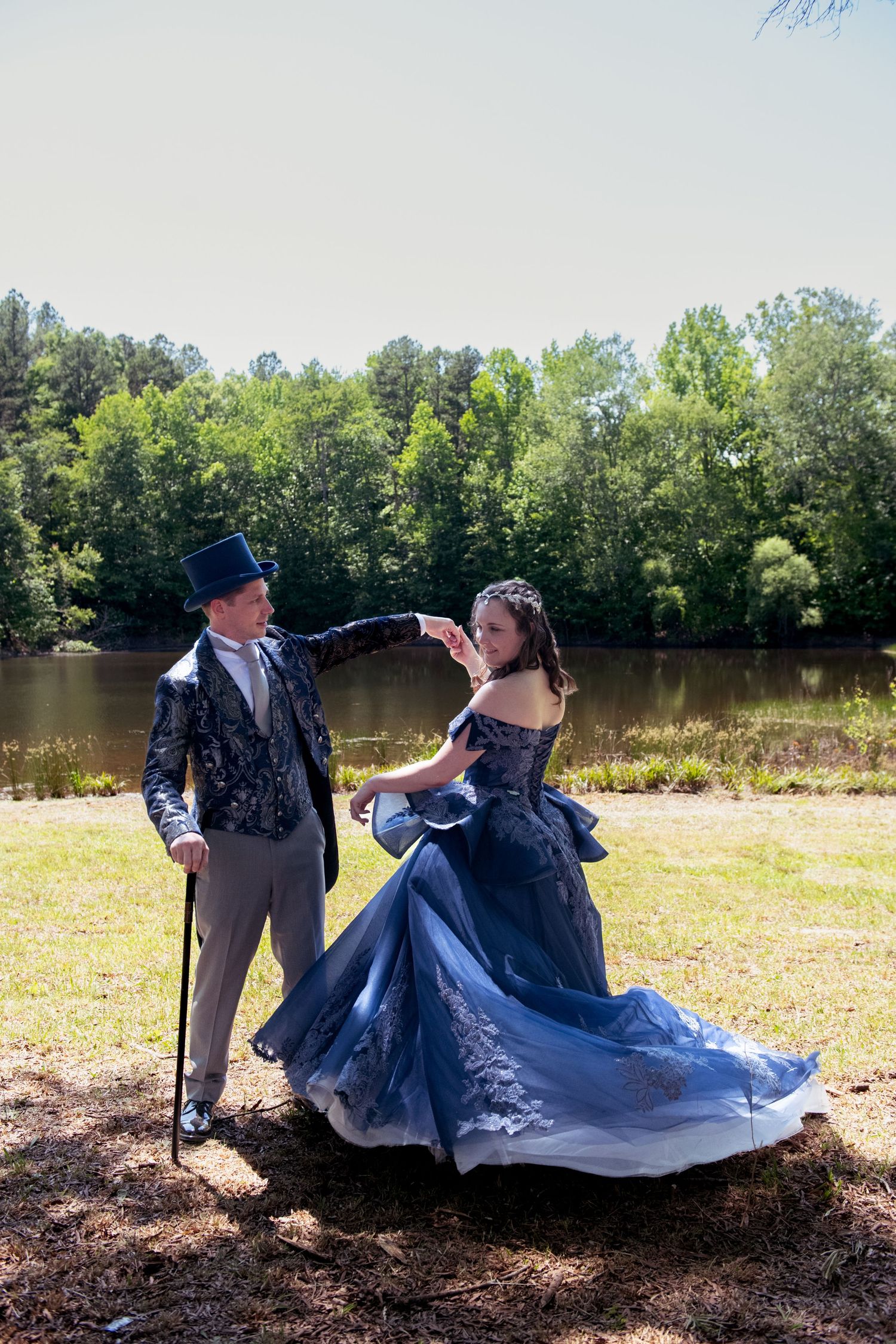Elegant couple dancing outdoors in vintage formal wear, blue gown and top hat, near a serene lake.