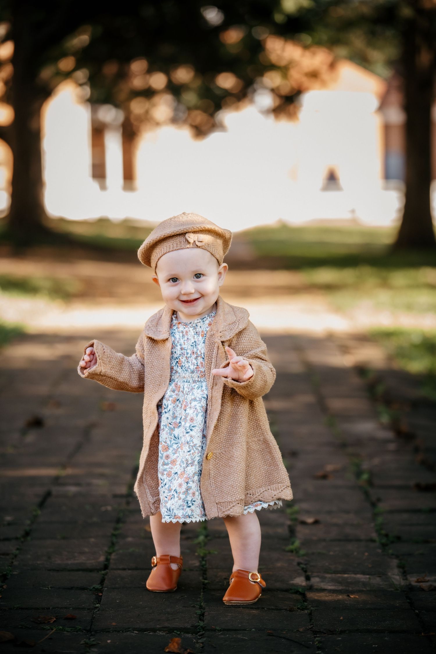 Smiling toddler girl in a tan coat, floral dress, and beret hat standing on a brick path in a sunny park.