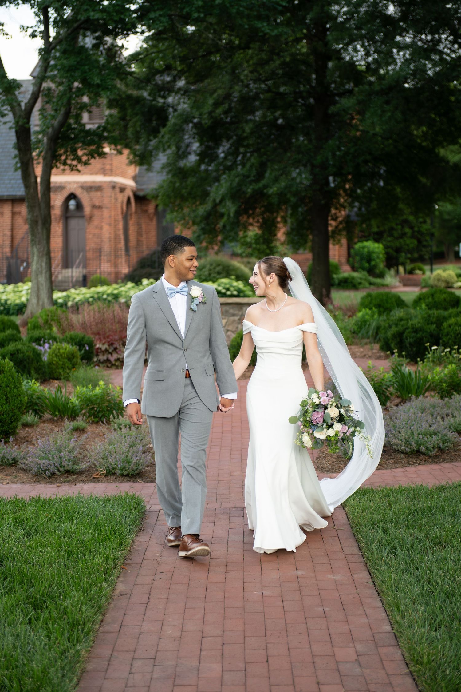 A bride and groom walk hand-in-hand along a brick garden path, surrounded by lush greenery and a historic brick building.