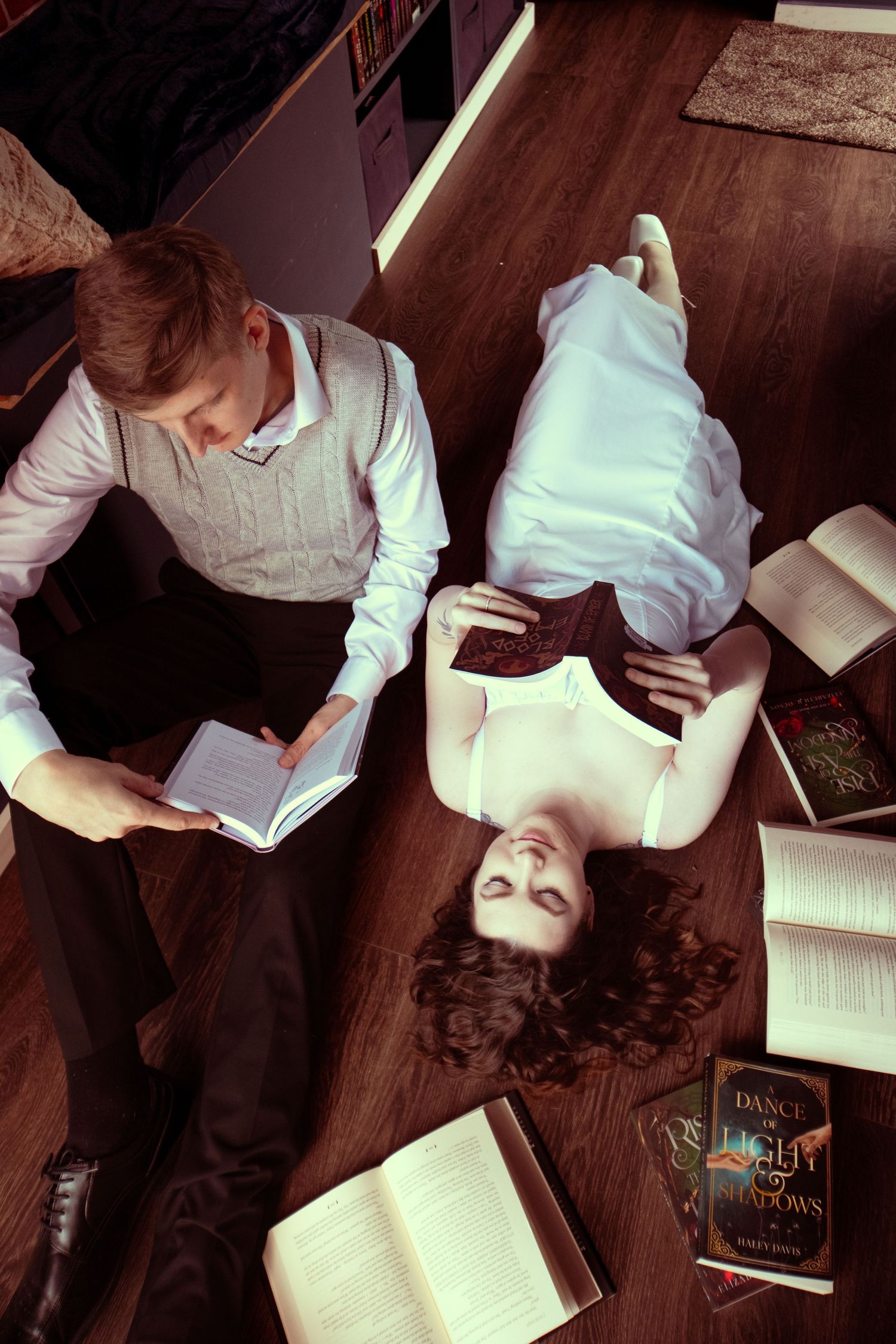 Man and woman relaxing on hardwood floor surrounded by open books in a cozy library setting.