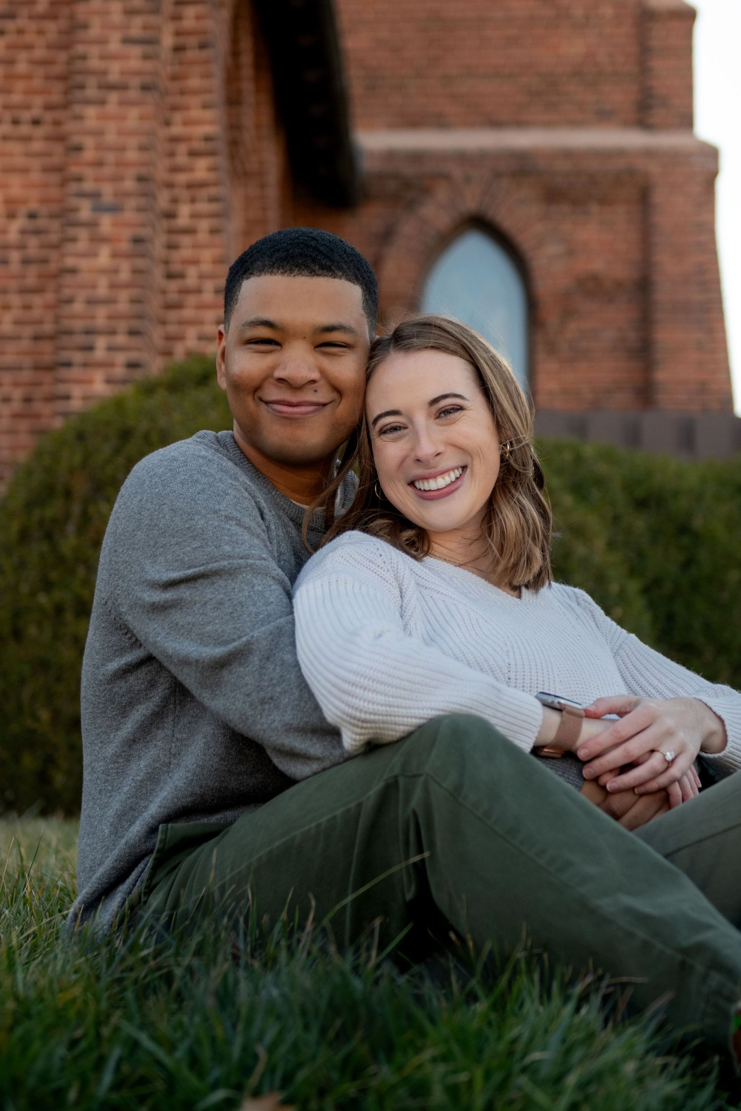 A couple sits together on grass in front of a brick building on a sunny day.