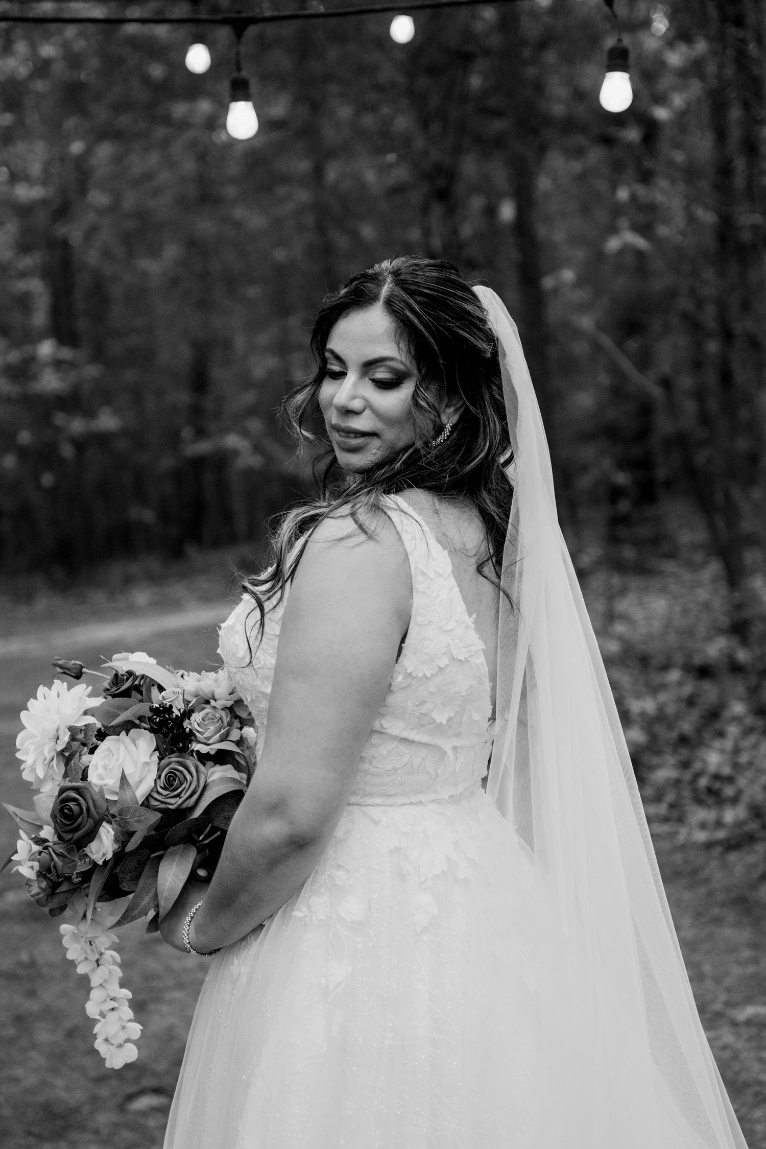 A bride in a white wedding dress and veil poses with her bouquet in a wooded outdoor setting.