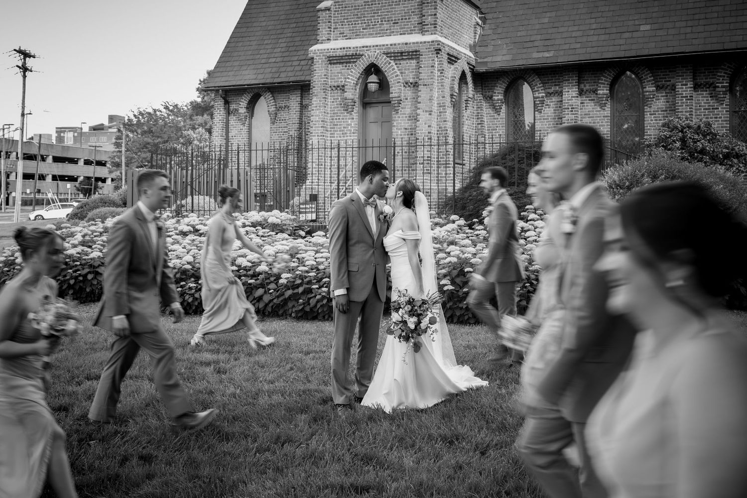 Black and white photo of bride and groom standing outside a brick church surrounded by wedding party guests.