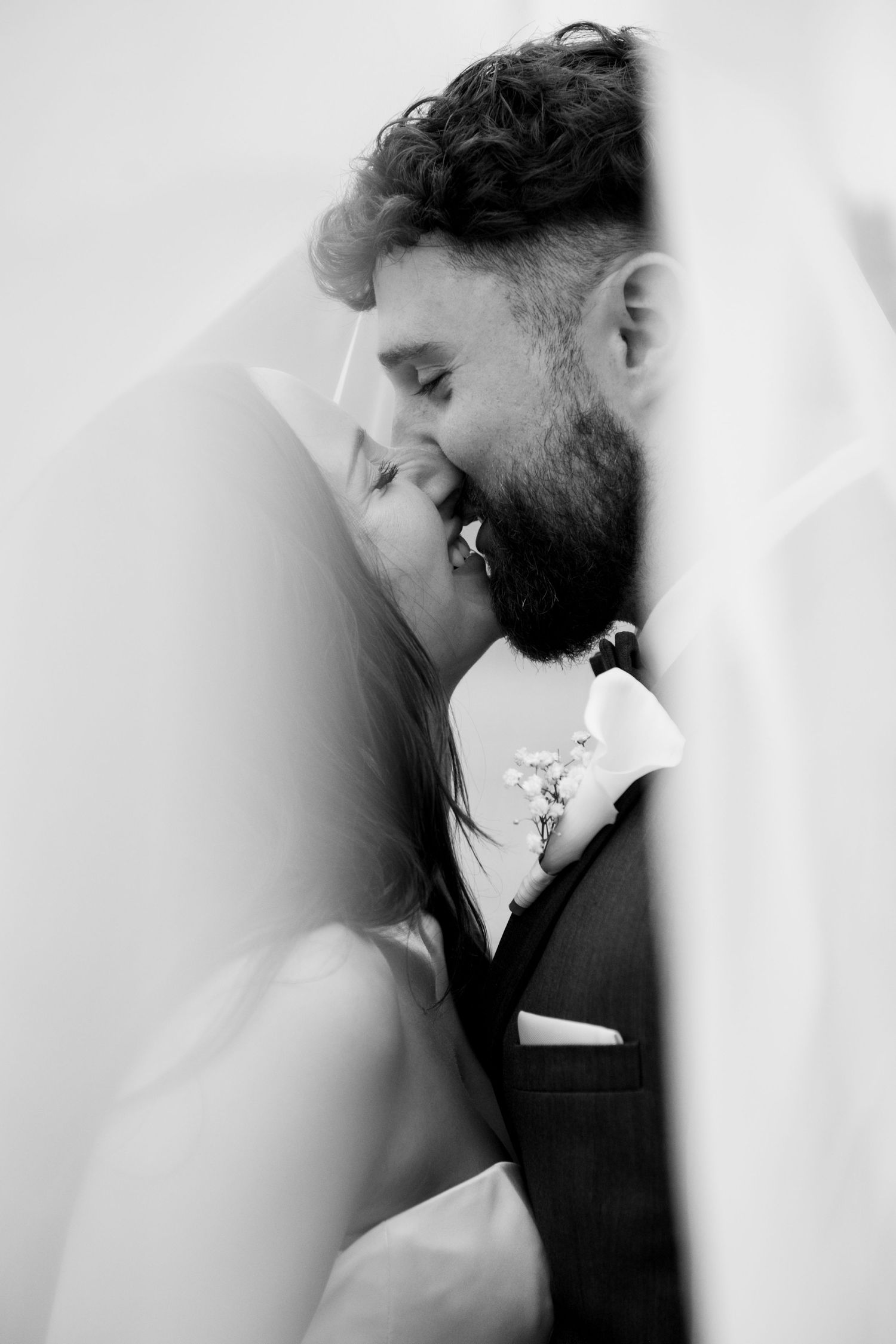 Black and white photo of a bride and groom sharing a romantic kiss under a flowing veil on their wedding day.