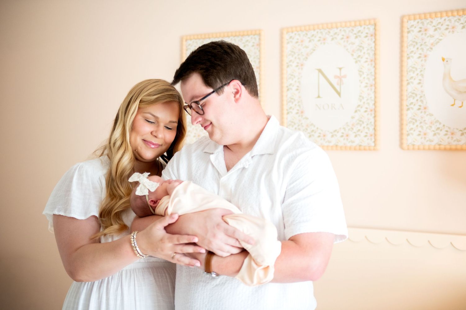 A couple in white clothing tenderly holds their newborn baby in a softly decorated nursery with framed letters on the wall.