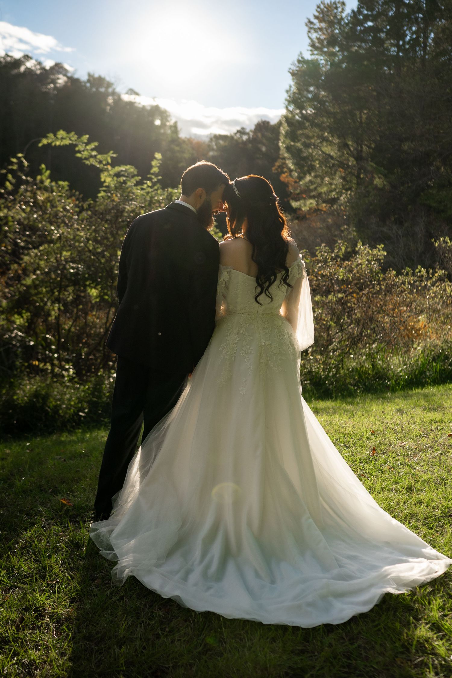 Bride in a flowing white lace gown and groom in a dark suit embrace foreheads together on a sunlit grassy lawn at an outdoor wedding.