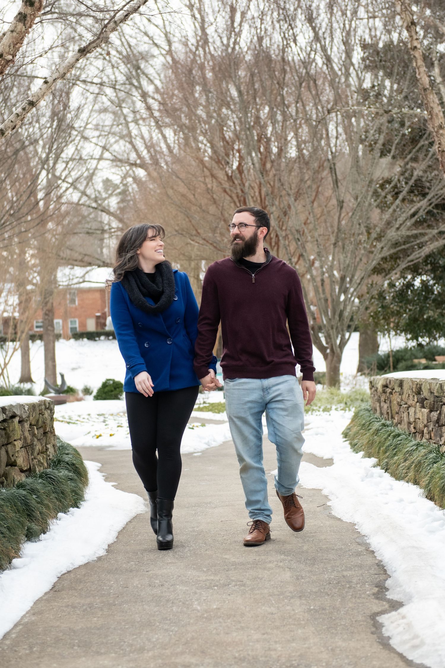A couple walks together on a snowy path through bare winter trees wearing blue and burgundy sweaters.