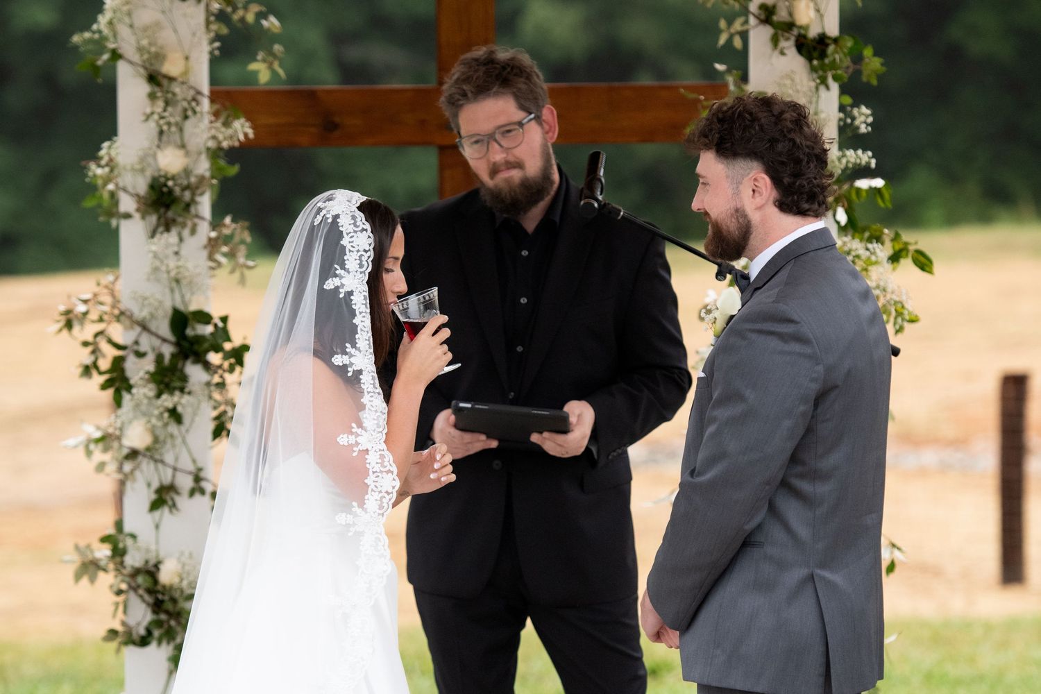 Bride and groom exchange vows outdoors at rustic wedding ceremony with officiant and wooden cross backdrop.