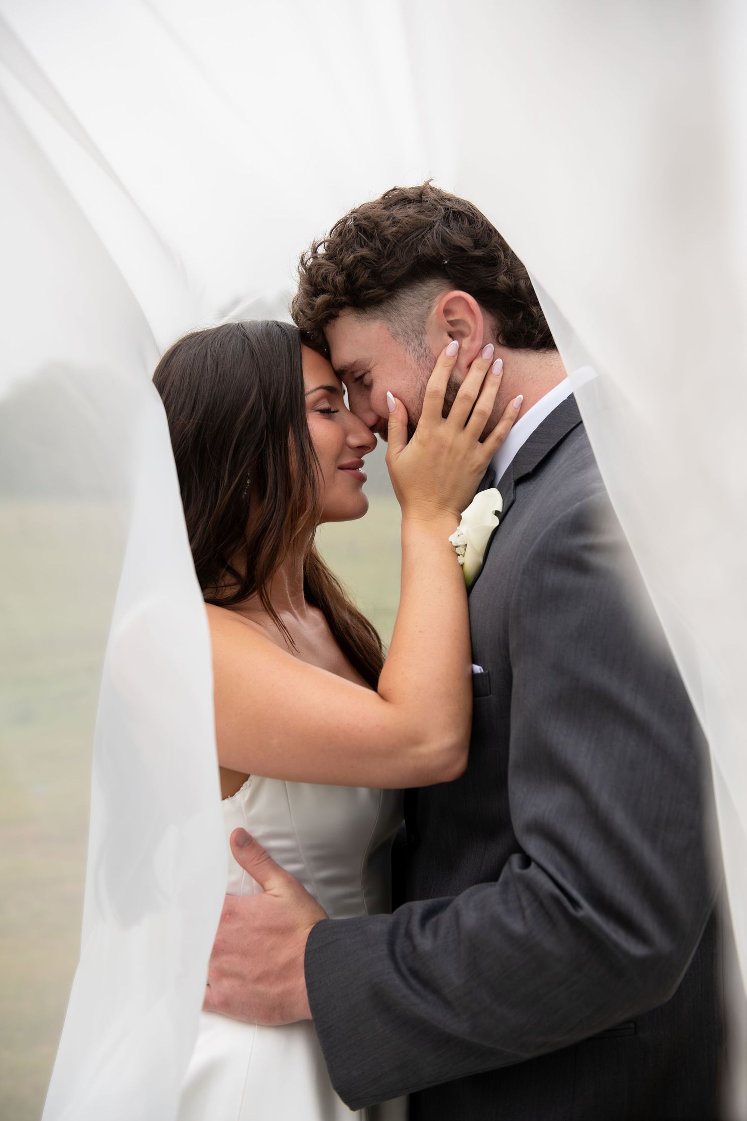 Bride and groom share a romantic moment under a white veil, with the bride tenderly holding the groom's face close to hers.