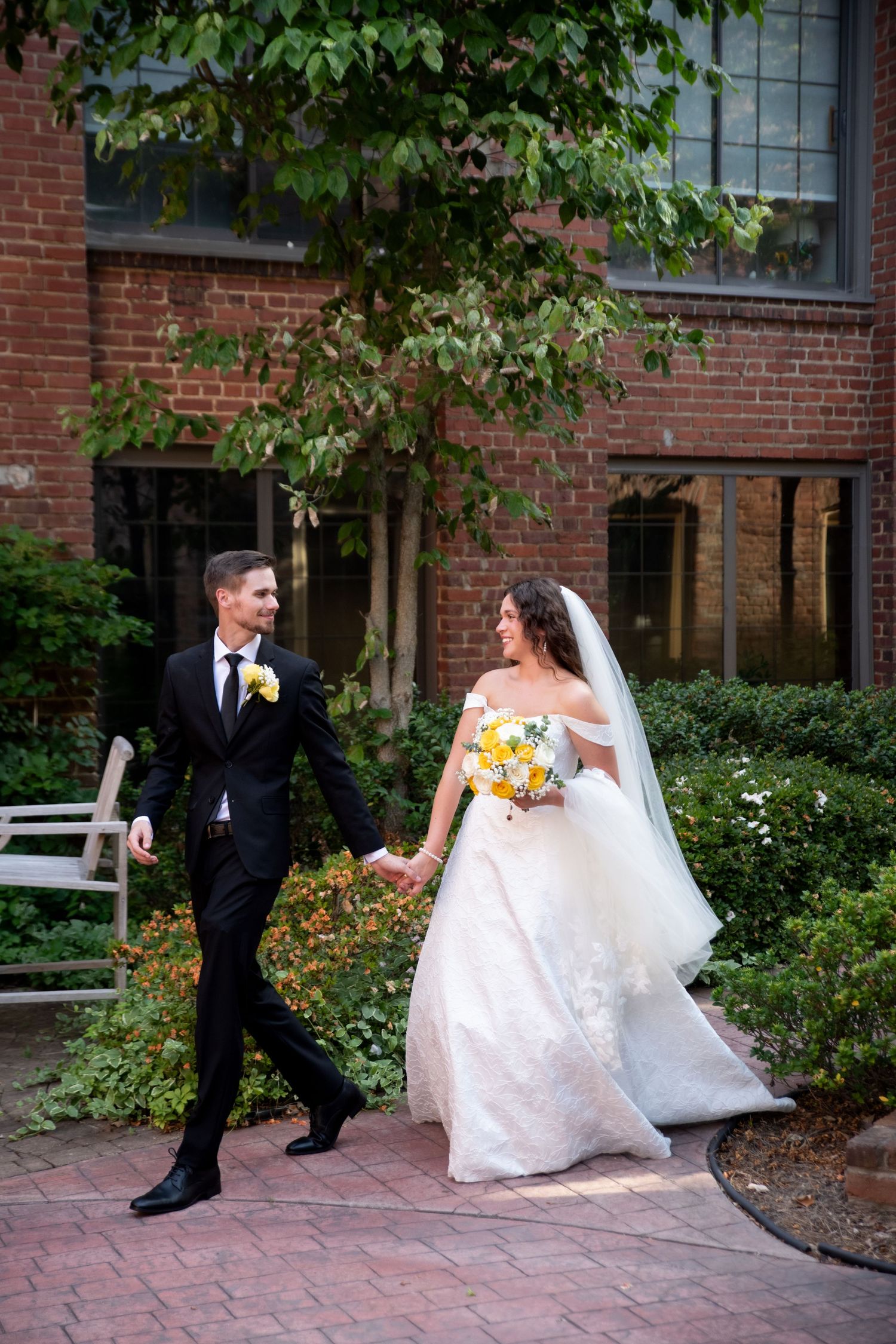 A bride and groom walk hand-in-hand on a brick path outside a red brick building, she holds a yellow bouquet.