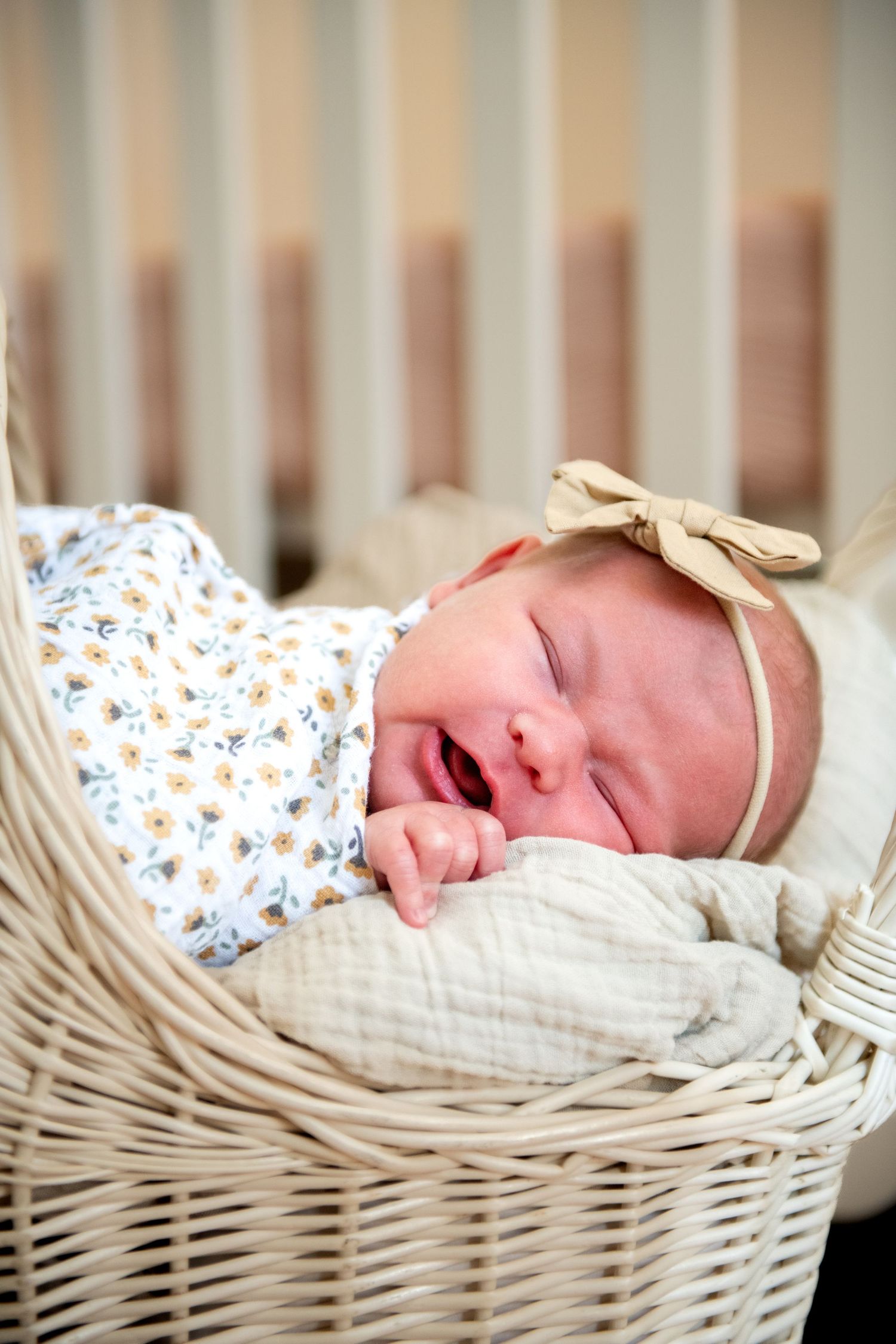 Newborn baby girl in floral swaddle and bow headband sleeping in a white wicker bassinet.