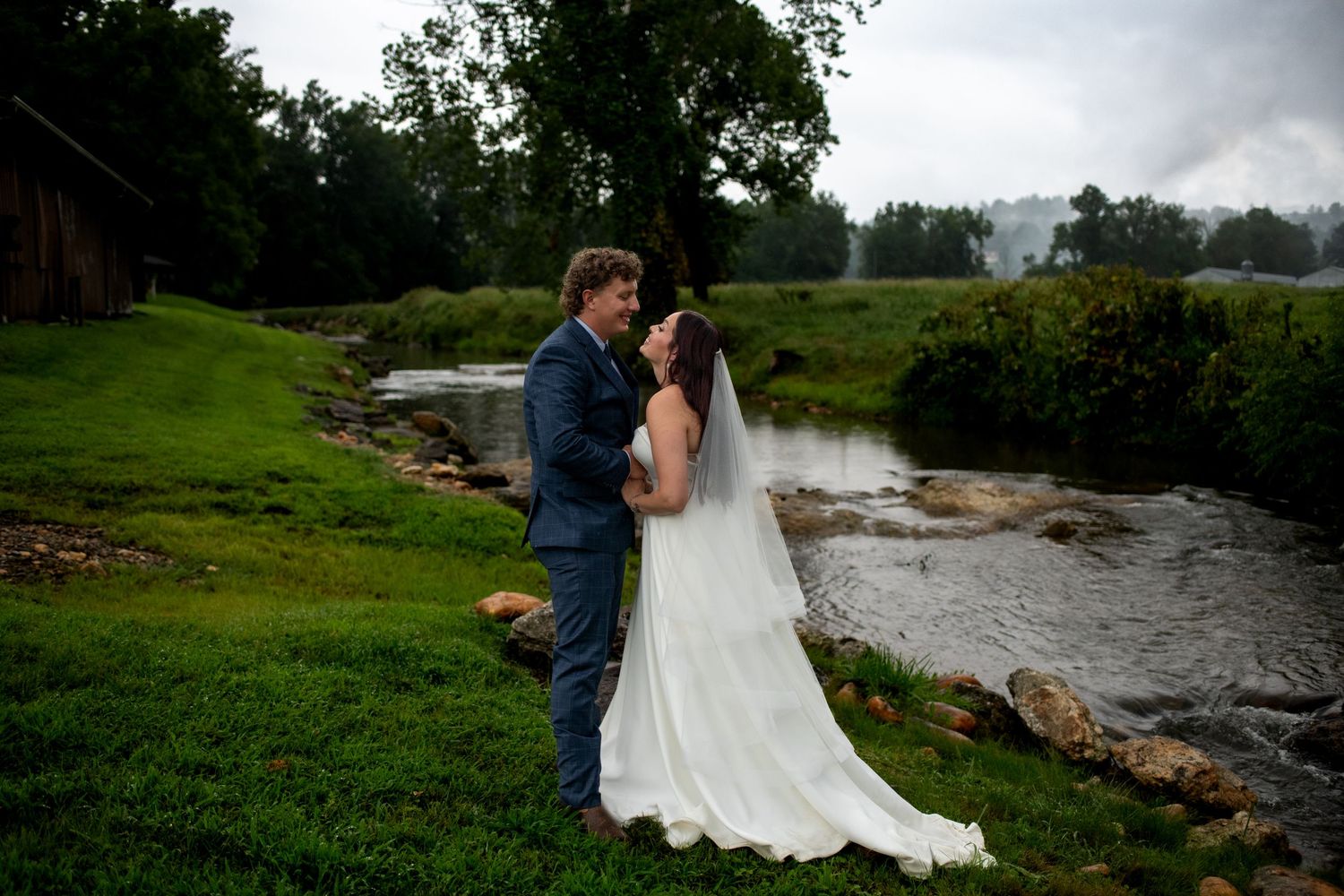 A wedding couple stands together near a flowing stream on a lush green landscape.