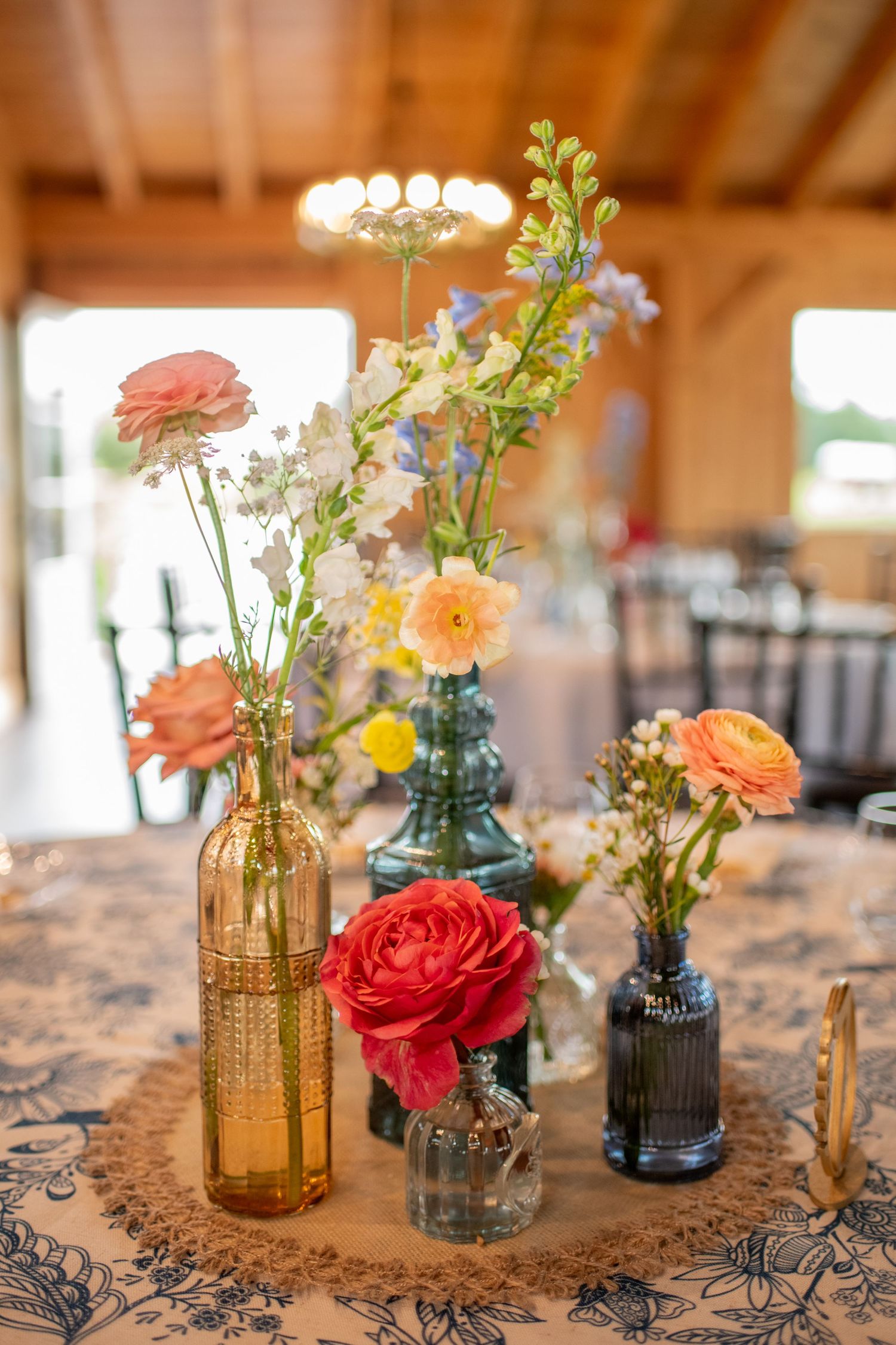 Vintage-style flower arrangements in colored glass vases on a patterned tablecloth.