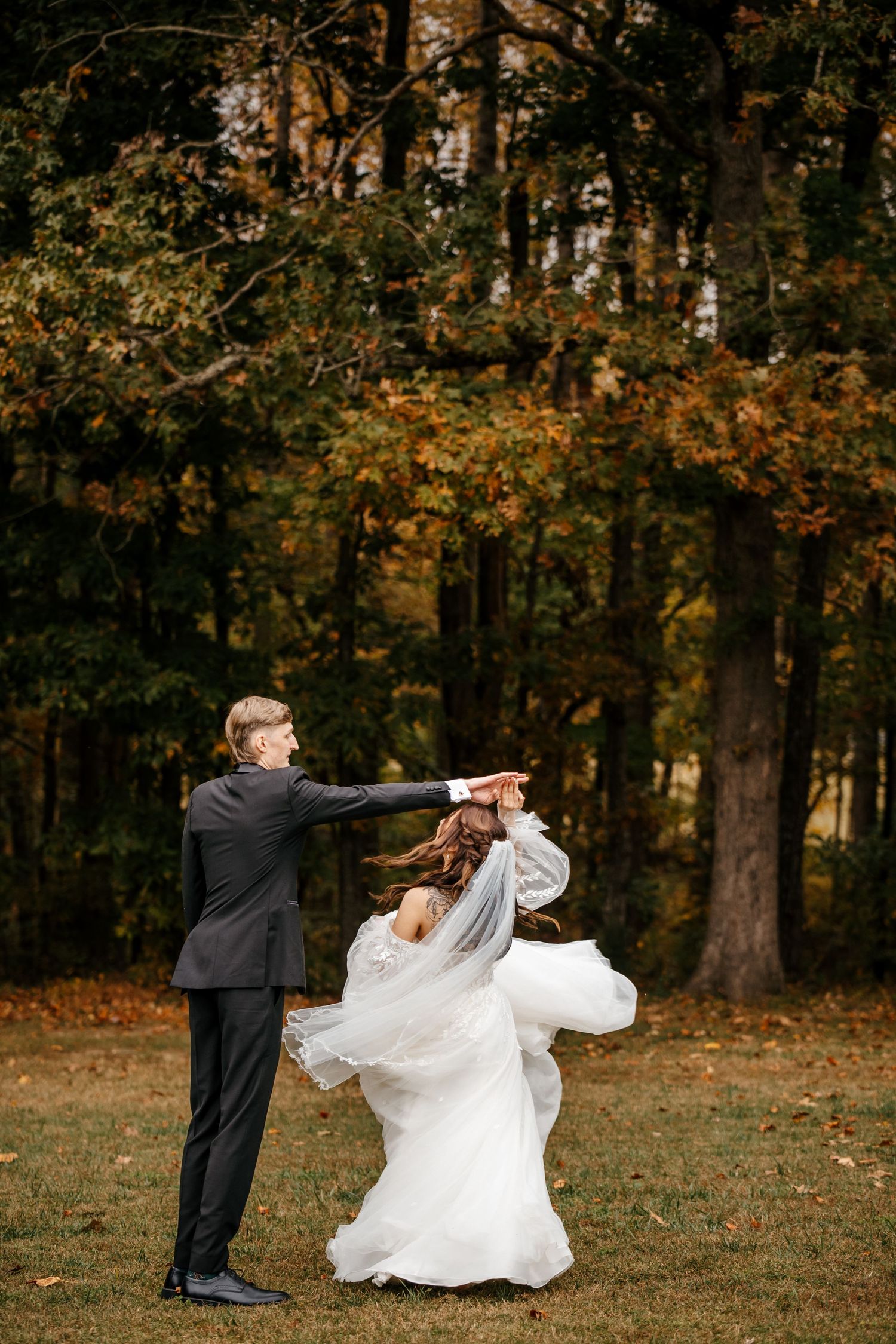 Groom in black suit twirling bride in white wedding gown outdoors in autumn forest setting.