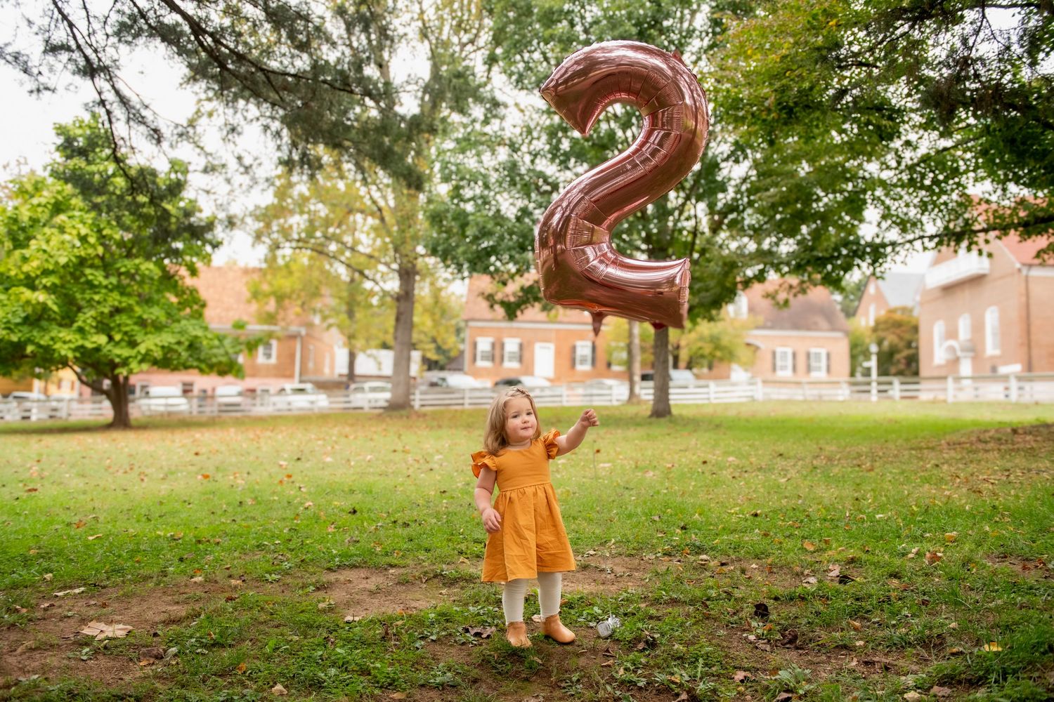 Toddler girl in yellow dress holding a number 2 balloon in a park, celebrating her 2nd birthday outdoors.